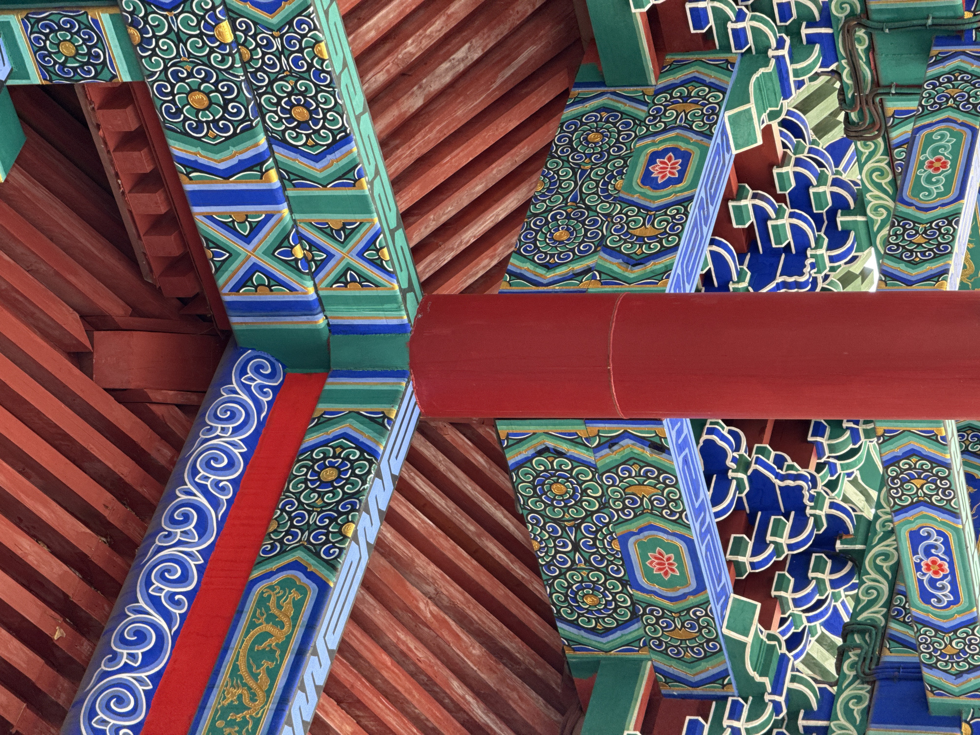 The image shows the ornately decorated interior ceiling of a pavilion with a red central column. The ceiling features intricate patterns in vibrant colors, including red, blue, green, and gold. These patterns are applied to wooden beams and architectural elements, creating a visually rich and traditional aesthetic. The red wooden beams of the roof are visible above the decorated structures. The scene appears to be an architectural detail within a larger structure, likely a temple or traditional building. There are no people visible in the image.
This image was taken at the "Smile of Khokhloma" pavilion at the VDNKh (Exhibition of Achievements of National Economy) in Moscow, Russia.
