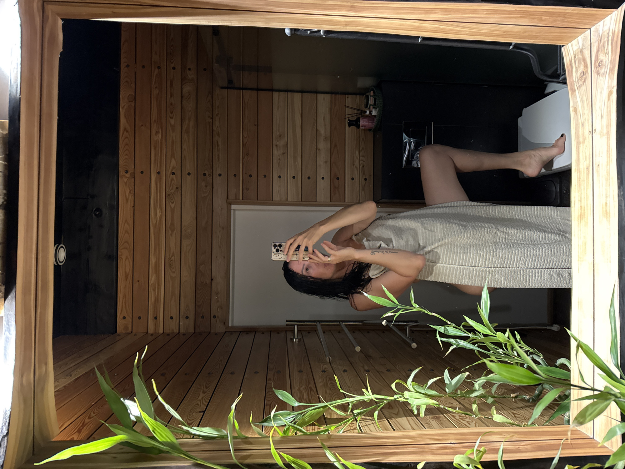 In a bathroom with wooden walls and flooring, a person is taking a selfie in a mirror. The person is wrapped in a light-colored towel and is holding up a smartphone. Their legs are extended, with one bent at the knee. A toilet is visible in the background. Green bamboo-like plants are in the foreground, with sunlight casting shadows. The overall impression is of a private, possibly spa-like setting. The image is a reflection, so the orientation is reversed.