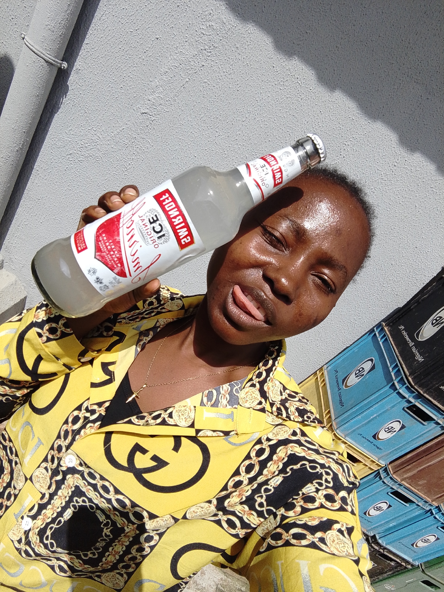 The image shows a young Black woman holding a Smirnoff Ice bottle and posing for a selfie outdoors. She is wearing a yellow shirt with a black and gold chain print, and a gold necklace with a cross pendant. Her tongue is sticking out playfully. Behind her, several stacked crates are visible, some blue and some brown, with branding that includes the initials "NB". The background appears to be a textured, light-colored wall, possibly painted concrete, with a pipe running vertically on the left. The scene suggests a casual outdoor setting, potentially in a residential or commercial area of Jos, Nigeria, given the presence of the crates which could be for beverages. The lighting indicates it is daytime, with sunlight casting shadows.