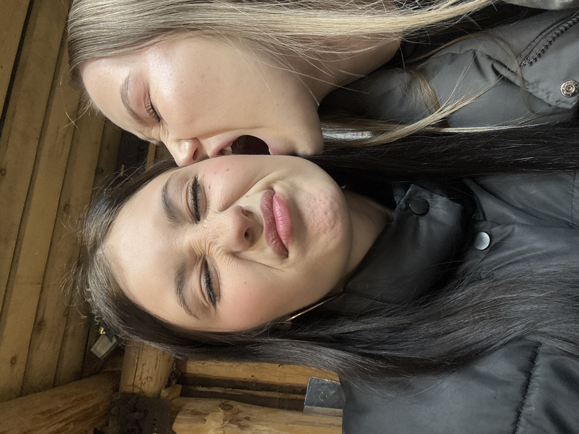 Two women lie side-by-side. The woman on top, with blonde hair, is playfully attempting to bite the ear of the woman beneath her, who has dark hair and is grimacing. Both are wearing dark-colored, puffy jackets. The background appears to be wooden, possibly part of a rustic structure. The location is not discernible as Фоки, Russia, from the visual information provided.
