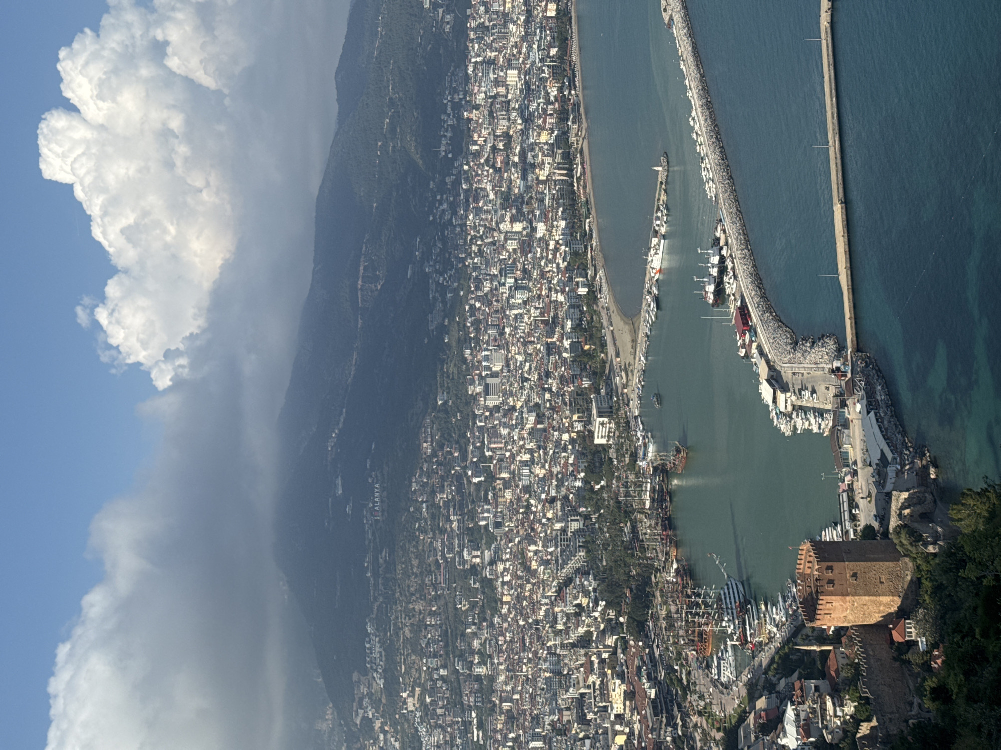 This aerial view captures a vibrant coastal city nestled at the base of lush green mountains, extending down to a bustling harbor and the sparkling blue-green sea. The sky above is partly cloudy, with large, puffy white clouds contrasting against a clear blue patch, suggesting daytime, likely late morning or early afternoon.
The cityscape is densely packed with numerous low-rise buildings, predominantly white or light-colored, with some featuring red-tiled roofs, sprawling across the mountain slopes and along the coastline. On the side of one of the mountains, large white letters clearly spell out "I ♥ ALANYA," indicating the location as Alanya, Turkey.
The harbor is a focal point, protected by a long, curving, rock-filled breakwater where numerous boats and yachts are moored. Further out, a very long, narrow concrete pier stretches into the open sea. Within the inner harbor, more boats, including larger tourist vessels with multiple masts, are visible.
Prominently featured at the entrance to the main harbor area is a distinctive reddish-brown octagonal stone tower, resembling a historic fortress or watchtower, perched on a rocky outcrop. Stone walls snake around the base of this tower, further suggesting a historical fortified area within the city. The clear water reveals the seabed in shallower parts of the harbor. The scene depicts a lively Mediterranean port city, likely a popular tourist destination.