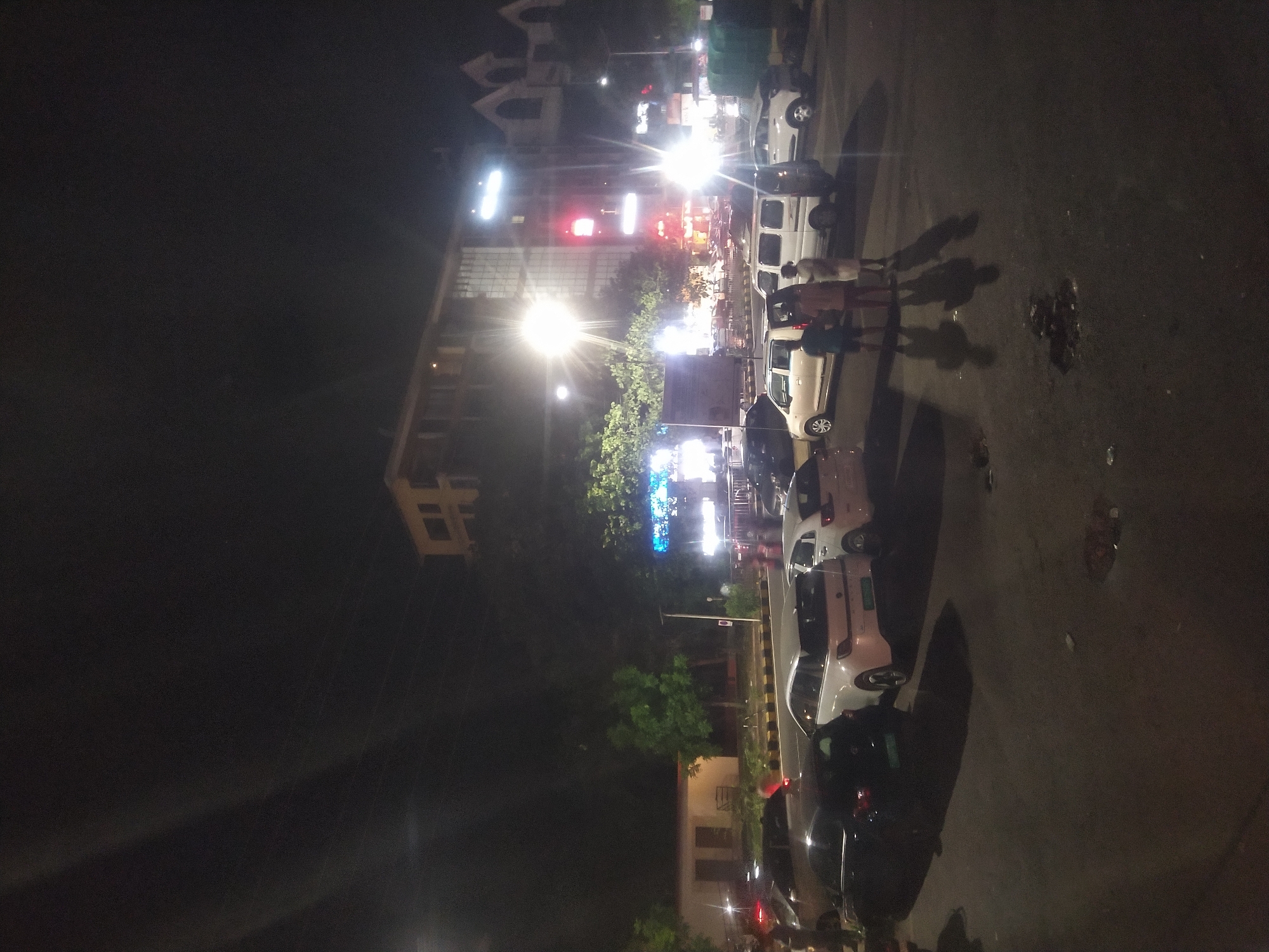 The image shows a street scene at night in Margao, India. Cars are parked and moving along the side of the road. A group of people are standing on the sidewalk with cars parked nearby. In the background, buildings are visible, illuminated by street lights and neon signs. A traffic light can also be seen. The overall lighting is low, with light sources creating strong contrasts and shadows. The road surface appears to be paved.