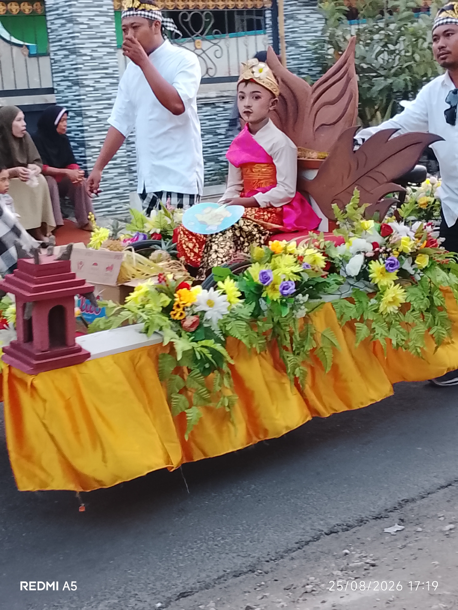 In Semanding, Indonesia, a parade float is adorned with yellow fabric, artificial flowers, and a decorative red structure. A young person in elaborate traditional attire, including a gold headdress and makeup, sits on the float. Next to them stands an adult male in traditional clothing. Other individuals, some in headscarves, are visible in the background, observing the procession. The scene appears to be taking place outdoors on a street.