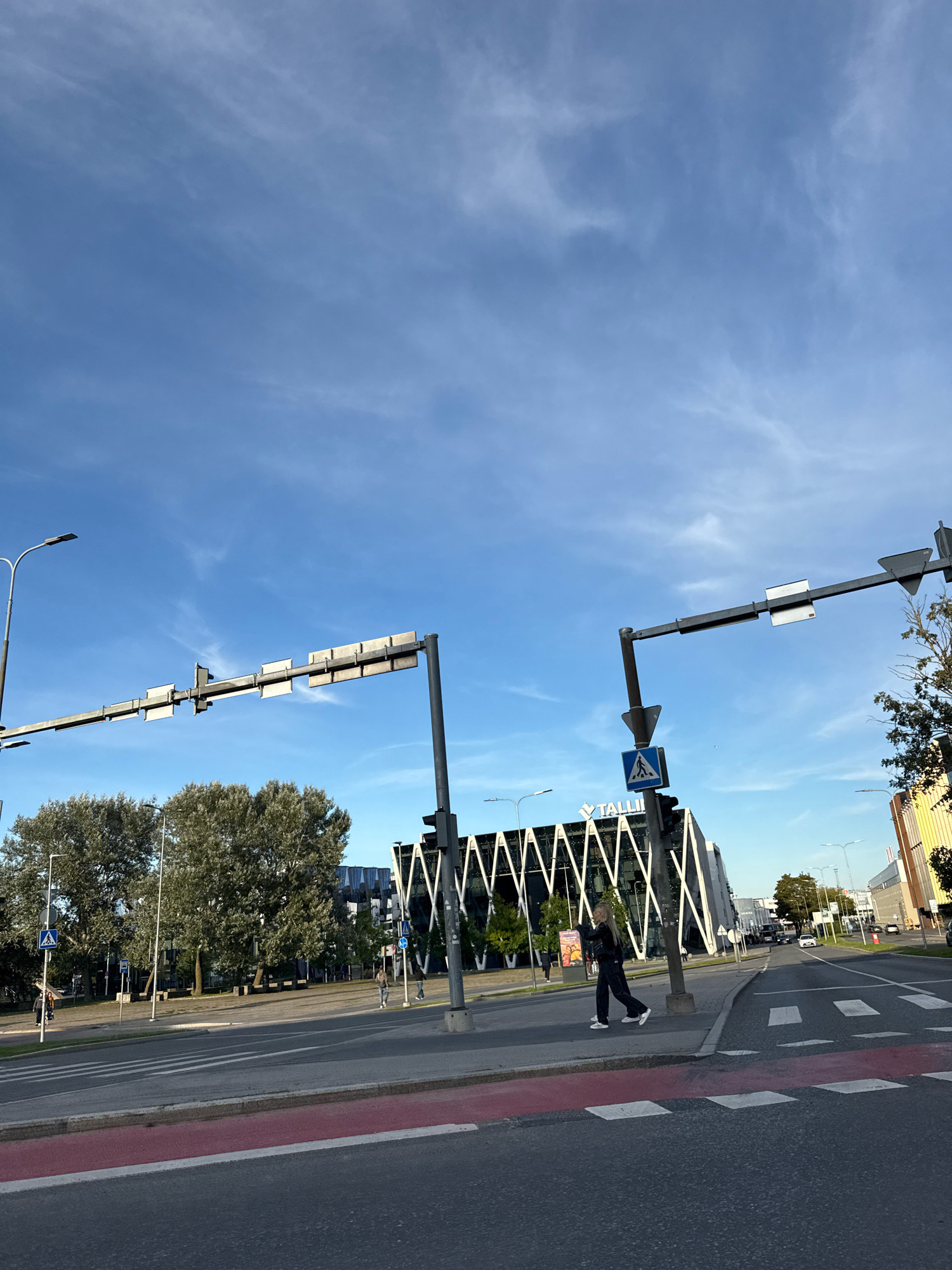 The image depicts a wide, paved road in Tallinn, Estonia, with a pedestrian crossing in the foreground. A person is walking across the pedestrian crossing from right to left. Behind the pedestrian crossing, there is a sidewalk lined with trees. Further back, a modern building with a distinctive angular architectural design and the word "TALLINN" is visible. Traffic lights and street lamps are present on both sides of the road. The sky is blue with some wispy clouds. In the background, other buildings and a few cars are visible. The scene appears to be a street in a commercial or urban area.