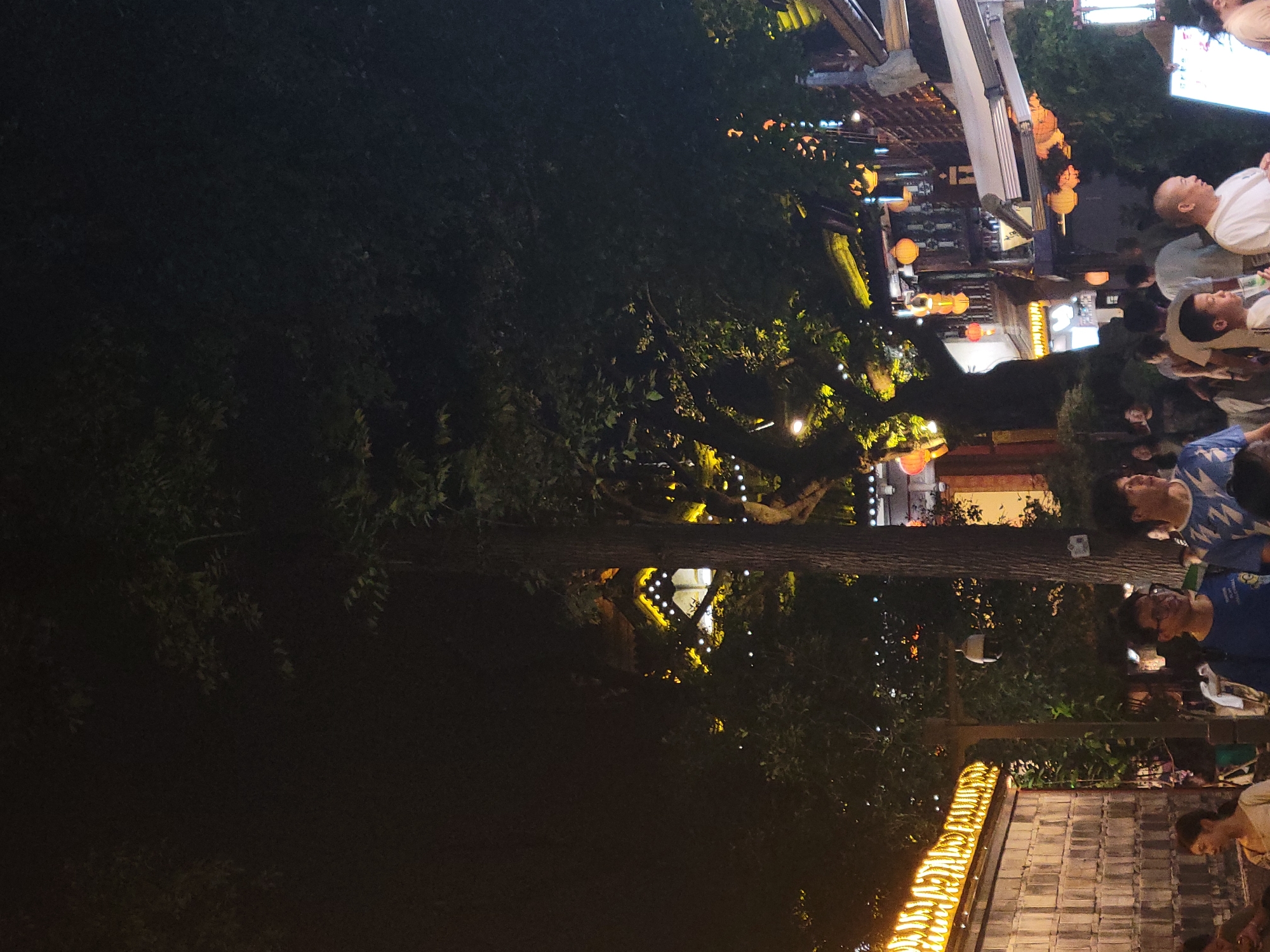 The image captures a nighttime scene in Wuhou District, China, featuring a street lined with traditional architecture and illuminated by various lights. A group of people are walking along the street, some are looking at something off-camera, and others are engaged in conversation. The architecture displays ornate details with yellow and white lights accentuating the eaves and windows. Red and yellow lanterns hang from the buildings and trees, adding to the festive atmosphere. Dense foliage from large trees partially obscures the view of the buildings and sky, casting shadows on the scene. The ground is paved, and there are visible street lamps and decorative lights. The overall impression is that of a bustling street during an evening event or gathering.