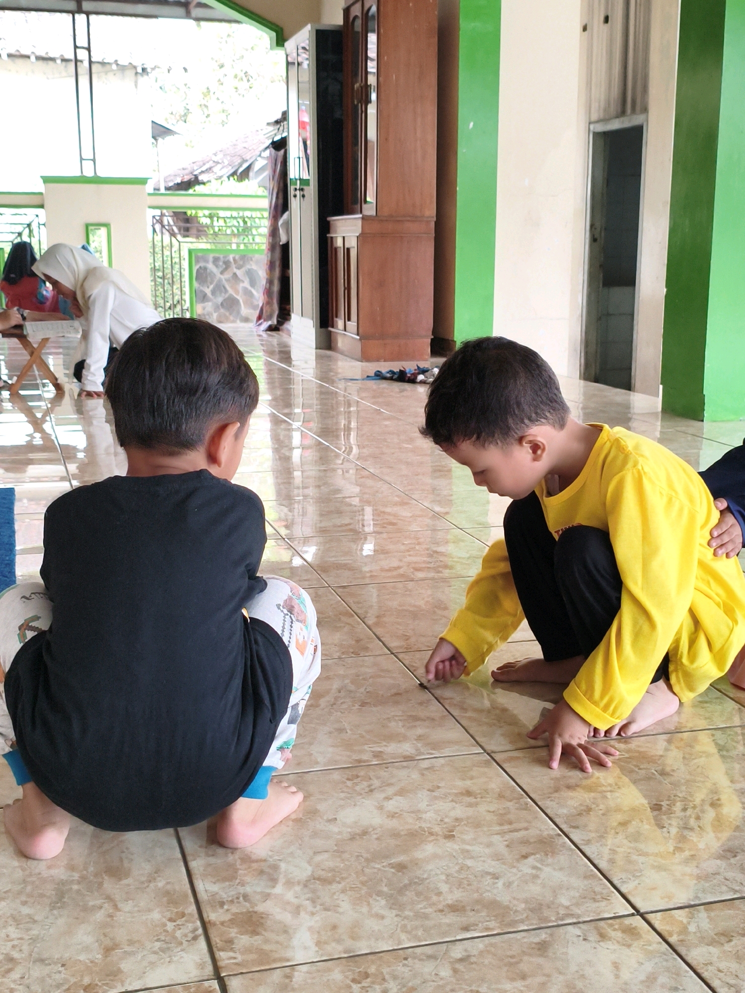 Two young boys are squatting on a tiled floor. The boy on the left wears a black t-shirt and patterned pants with blue trim. The boy on the right wears a yellow long-sleeved shirt and black pants. He is reaching down to touch something on the floor with his left hand. In the background, a woman wearing a white hijab is visible, as are wooden cabinets and green and cream-colored walls. The setting appears to be a communal space, possibly a mosque or community center, in Salatiga, Indonesia, suggested by the architecture and the presence of individuals in religious attire. The floor is shiny, reflecting the light and the figures.