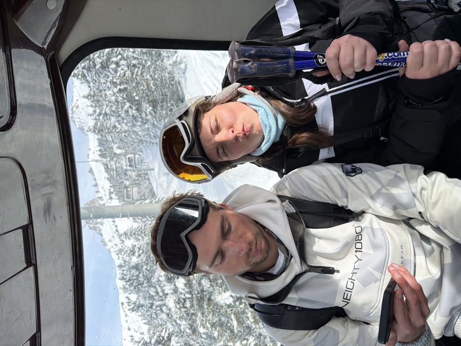Two individuals are seated inside a ski gondola cabin, traversing a snow-covered mountainous region in a ski resort near Les Allues, France. The male, positioned on the left, wears a white and grey patterned hooded jacket with "NEIGHTY 1080® GEAR" branding and black-framed ski goggles resting on his forehead. He is observed looking downwards at a black smartphone held in his left hand. A black backpack is visible on his shoulders. The female, positioned on the right, wears a black and white ski jacket, a light blue neck gaiter, and a grey ski helmet with orange-tinted goggles pushed up onto the helmet. She holds two blue and black ski poles, branded "IPRON" and "Ski-M.com", in her left hand and is making a puckered lip expression towards the viewer. Both individuals appear to be young adults. Through the gondola's window, the background reveals an extensive snow-covered landscape with dense coniferous trees and mountain slopes, under a clear sky, with a functional chairlift system visible in the distance.