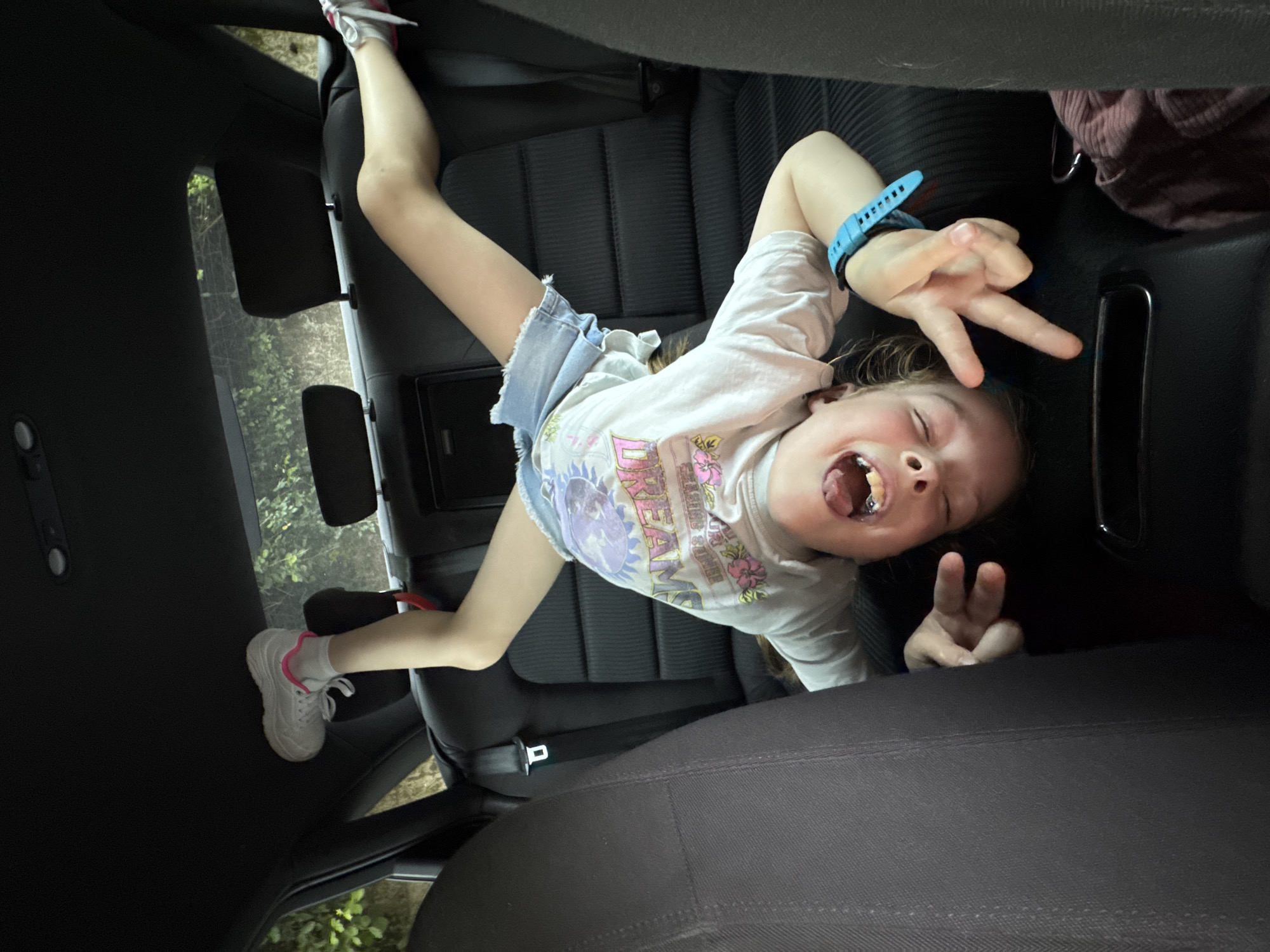 A child is lying in the back seat of a car. She is wearing shorts, a T-shirt and shoes. She is smiling and making the peace sign with both hands. The car is driving through a wooded area. The exact location in the city of Budva, Montenegro, is not discernable from the image.