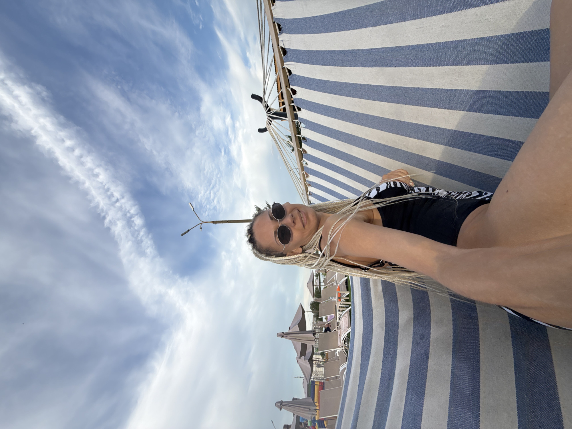 A woman with blonde braids and sunglasses is sitting on a beach blanket with blue and white stripes, in Rusanovo, Russia. She is wearing a black swimsuit. Behind her, beach chairs and umbrellas are visible under a cloudy sky.
