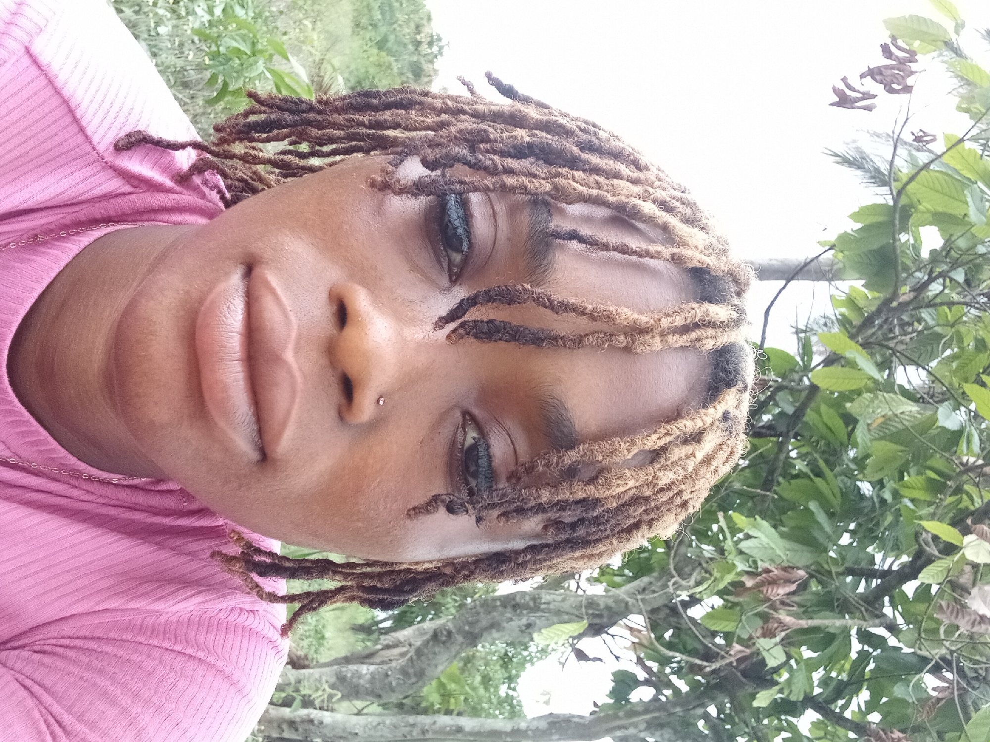 An overhead portrait-style shot captures a person with medium-brown skin and dreadlocks looking directly at the camera. The person's head is tilted slightly, and their expression is neutral. They have dark eyes and full lips. A small, clear or silver stud piercing is visible on the left side of their nose. Their dreadlocks are a mix of dark and lighter brown hues, with some strands falling across the forehead and around the face.
The person is wearing a pink, ribbed top, which features a horizontal striped texture. A thin gold chain is visible around their neck, resting on the fabric of the shirt. The background consists of lush green foliage and tree branches, indicating an outdoor setting, possibly in a garden or natural area. The sky in the upper right portion of the image is bright and appears overcast or hazy white. The lighting suggests it is daytime. The image hints at a location like Oba-Ile, Nigeria, given the natural outdoor environment. No visible text is present in the image.