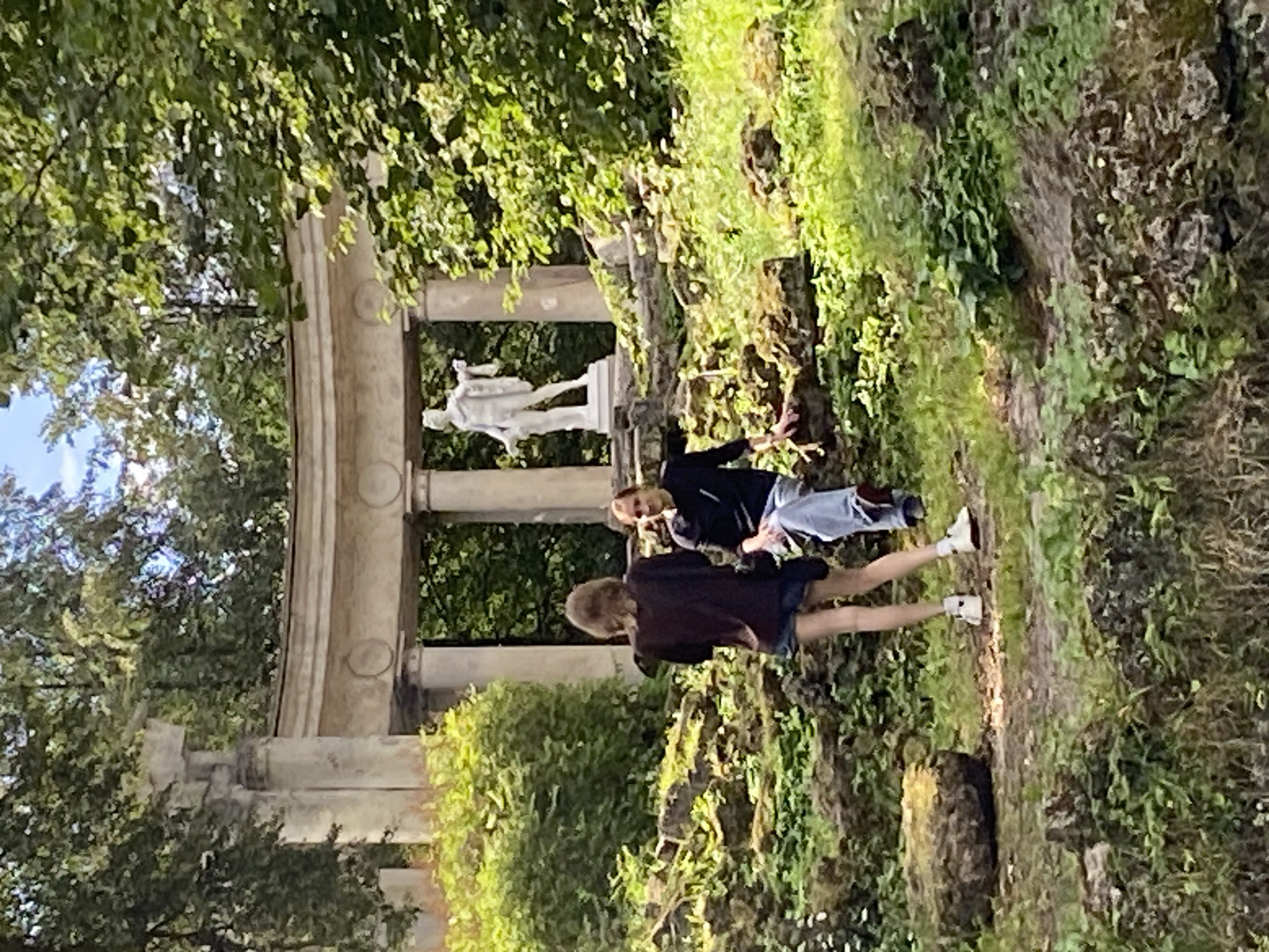In Pavlovsk, Russia, two individuals are observed in a park setting. A person with blonde hair, facing away from the camera, stands near a stone structure. This individual wears a dark top and denim shorts, with white sneakers. To their right, a second person, facing the camera, is seated. This individual has brown hair and wears a dark long-sleeved shirt with light-colored pants and shoes. They are positioned on a rocky, moss-covered incline. In the background, a white classical statue stands within an open-air colonnade, framed by lush green foliage and trees. The environment appears to be a well-maintained historical park with a mix of natural elements and architectural features.