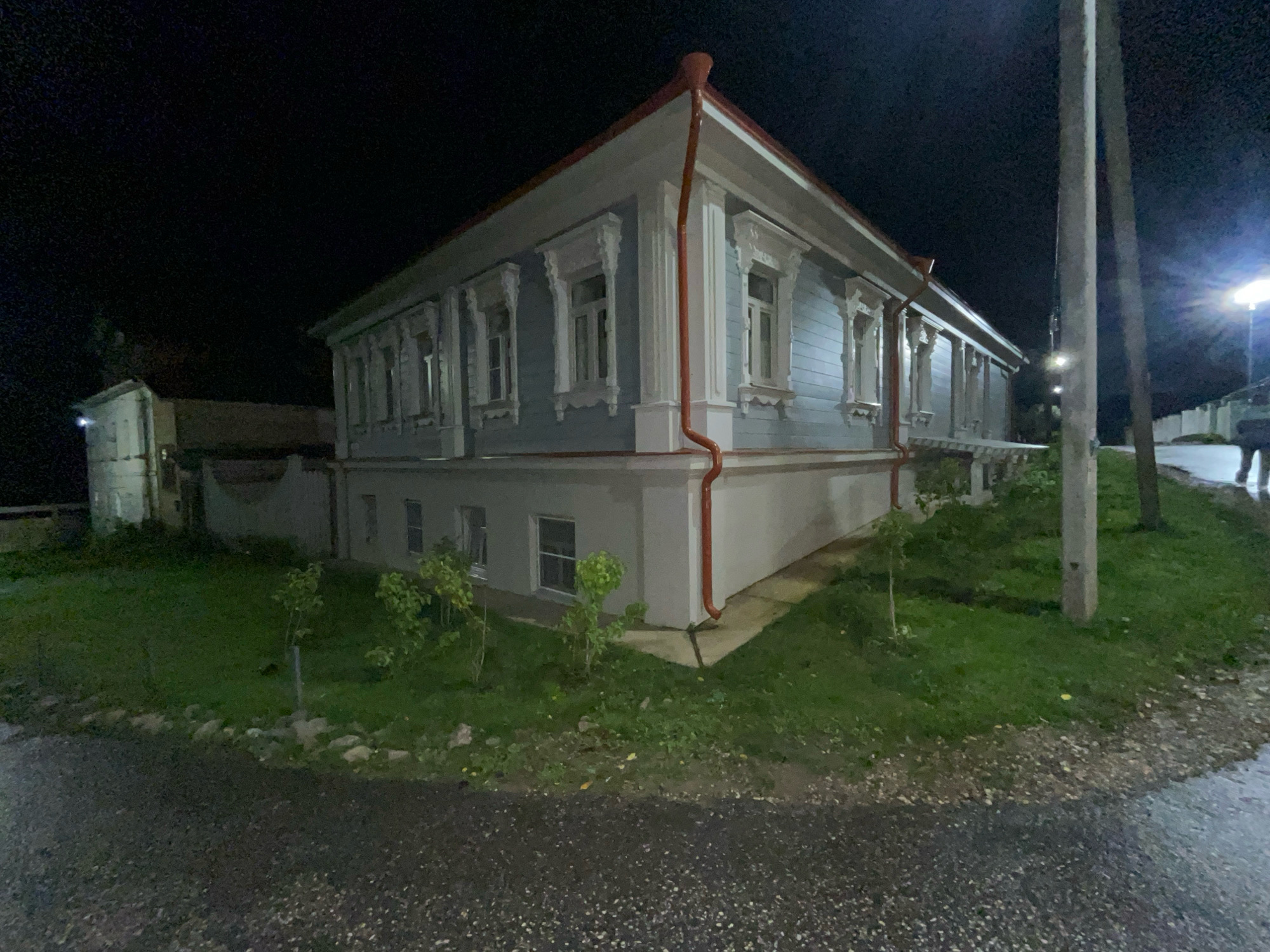 A nighttime exterior view shows a light blue wooden building with white trim and red gutters, likely a historical structure. The building has multiple windows with decorative white carvings around them. To the left of the main building, a smaller white structure is visible, possibly an annex or outbuilding. A concrete utility pole stands to the right of the main structure, with another pole behind it. The ground in front of the building is covered in green grass with small trees planted. A paved area, possibly a road or path, is in the foreground, appearing wet and reflective, suggesting recent rain. The sky is dark and clear. The scene appears to be in a quiet, possibly rural or suburban, setting at night. The architectural style suggests it could be in a historical area of Suzdal, Russia, known for its preserved wooden architecture. No people are clearly visible in the frame.