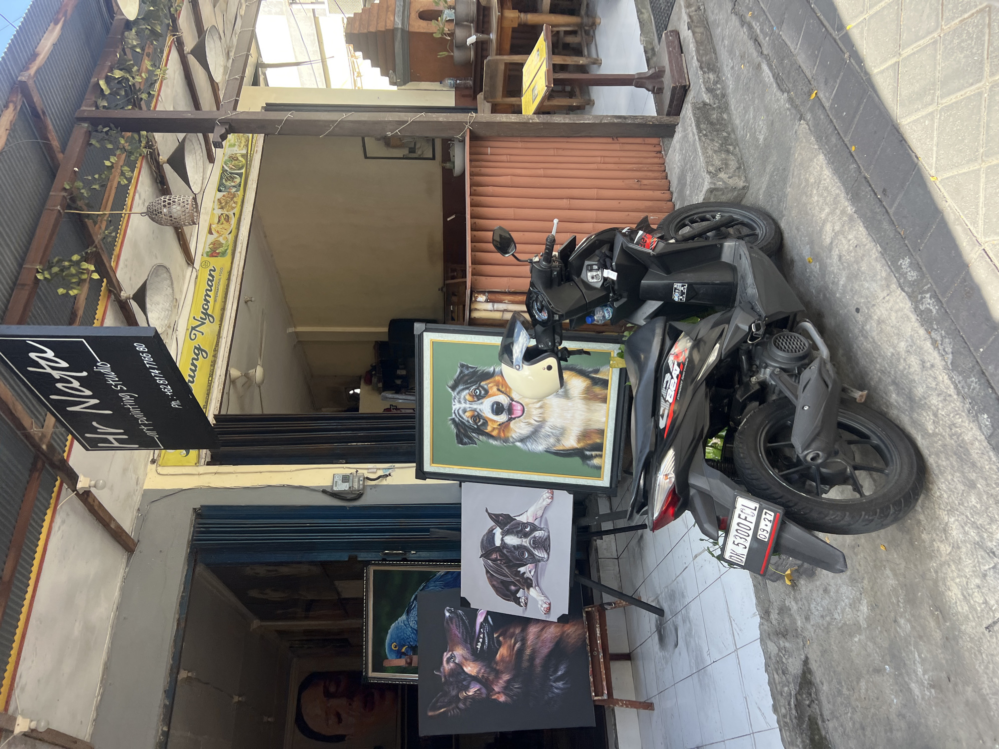 In Kuta, Indonesia, a black scooter is parked in front of an art painting studio and a restaurant. The studio, named "Hr Nata art painting studio," displays several dog portraits, including a German Shepherd and a Boston Terrier, and a blue parrot painting. A large painting of an Australian Shepherd is prominently displayed next to the scooter, with a yellow helmet resting on its frame. The restaurant, identified by a sign with "Warung Nyoman" and images of food, is visible behind the studio. The area appears to be a commercial street with tiled sidewalks and a corrugated metal roof overhanging the establishments. The scooter has a license plate that reads "DK 5300 FCL" and shows an expiration date of "09-27."