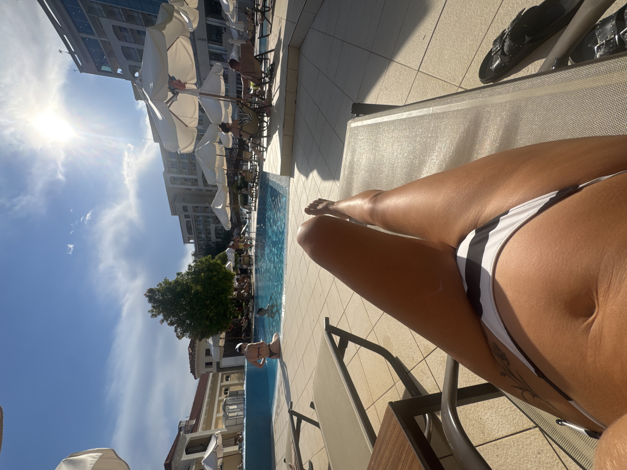 This image captures a sunny day at a resort pool in Boreti, Montenegro. In the foreground, a person's legs and torso are visible, lying on a lounge chair. They are wearing a black and white striped bikini. To the left of the foreground, a side table is present. In the mid-ground, a swimming pool is visible, with several people in and around it. A woman in a bikini is sitting on the edge of the pool, and another person is partially submerged in the water. Lounge chairs and umbrellas are scattered around the poolside, and other guests are seen relaxing. In the background, a multi-story hotel building stands against a bright blue sky with scattered clouds and bright sunlight. The environment is paved with light-colored tiles.