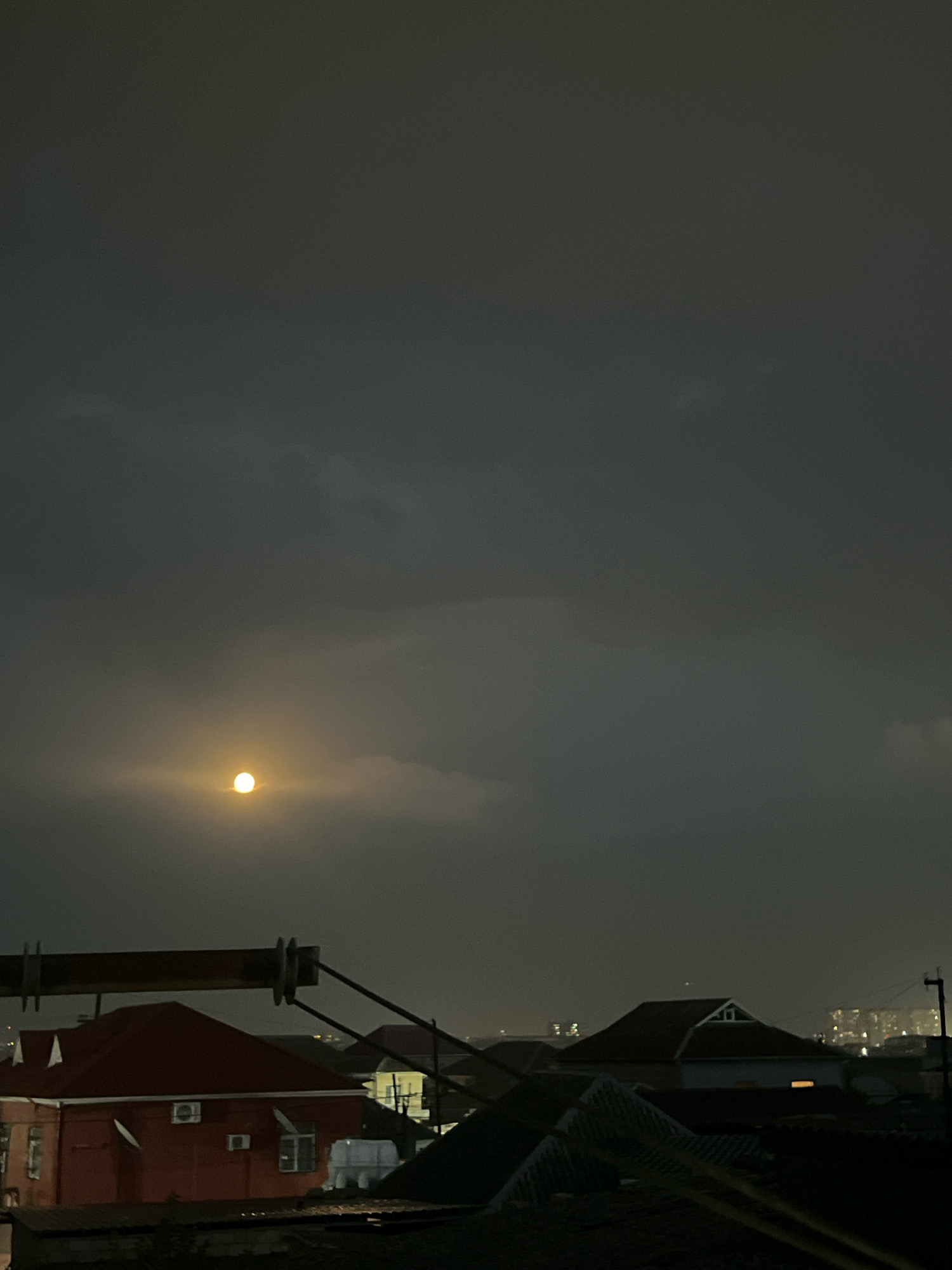 The image is a nocturnal, wide-angle shot taken from an elevated position, looking out over a residential area. The sky is dark and overcast, with a hazy, luminous circle suggesting the moon partially obscured by clouds, positioned slightly left of center. In the foreground, silhouetted against the faint sky, are the dark shapes of rooftops. Dominating the mid-ground is a red-brick building with visible air conditioning units and windows, and adjacent structures with red and brown tiled roofs. Further in the distance, more rooftops and buildings are visible, with some distant city lights suggesting a populated area. A metallic structure, possibly part of an overhead line or antenna, crosses the upper mid-section of the frame. The scene appears to be a view of houses and urban infrastructure in the Sabunchu district of Baku, Azerbaijan, at night. There are no people visible in the image.