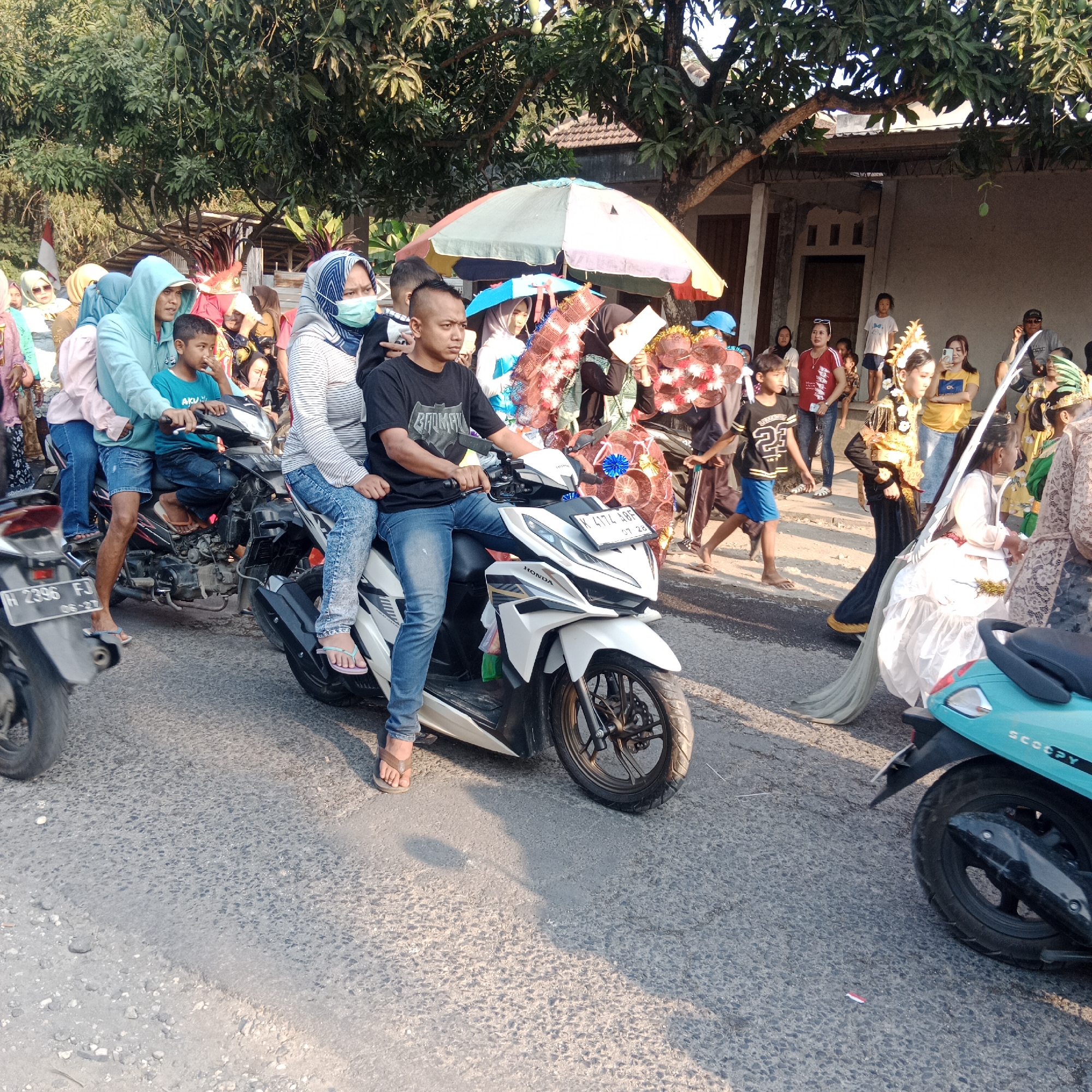 In a street scene in Tegalrejo, Indonesia, a procession is underway, possibly a parade or festival. A group of people, including men, women, and children, are gathered on both sides of the asphalt road. Several individuals are riding on motorbikes. In the foreground, a white motorbike is prominent, with a man driving and a child seated behind him. Another motorbike is visible on the left side of the image, and the rear of a teal scooter is seen on the right.
The procession appears to feature traditional elements, with some individuals dressed in elaborate costumes, including headdresses and flowing garments. Decorative items, such as colorful, possibly paper-made, decorations, are being carried or displayed. An umbrella, providing shade, is positioned above some of the participants. The background shows residential buildings with tiled roofs and a large tree with lush foliage, suggesting a warm, possibly tropical, climate. The overall atmosphere suggests a communal event with a celebratory or cultural significance.