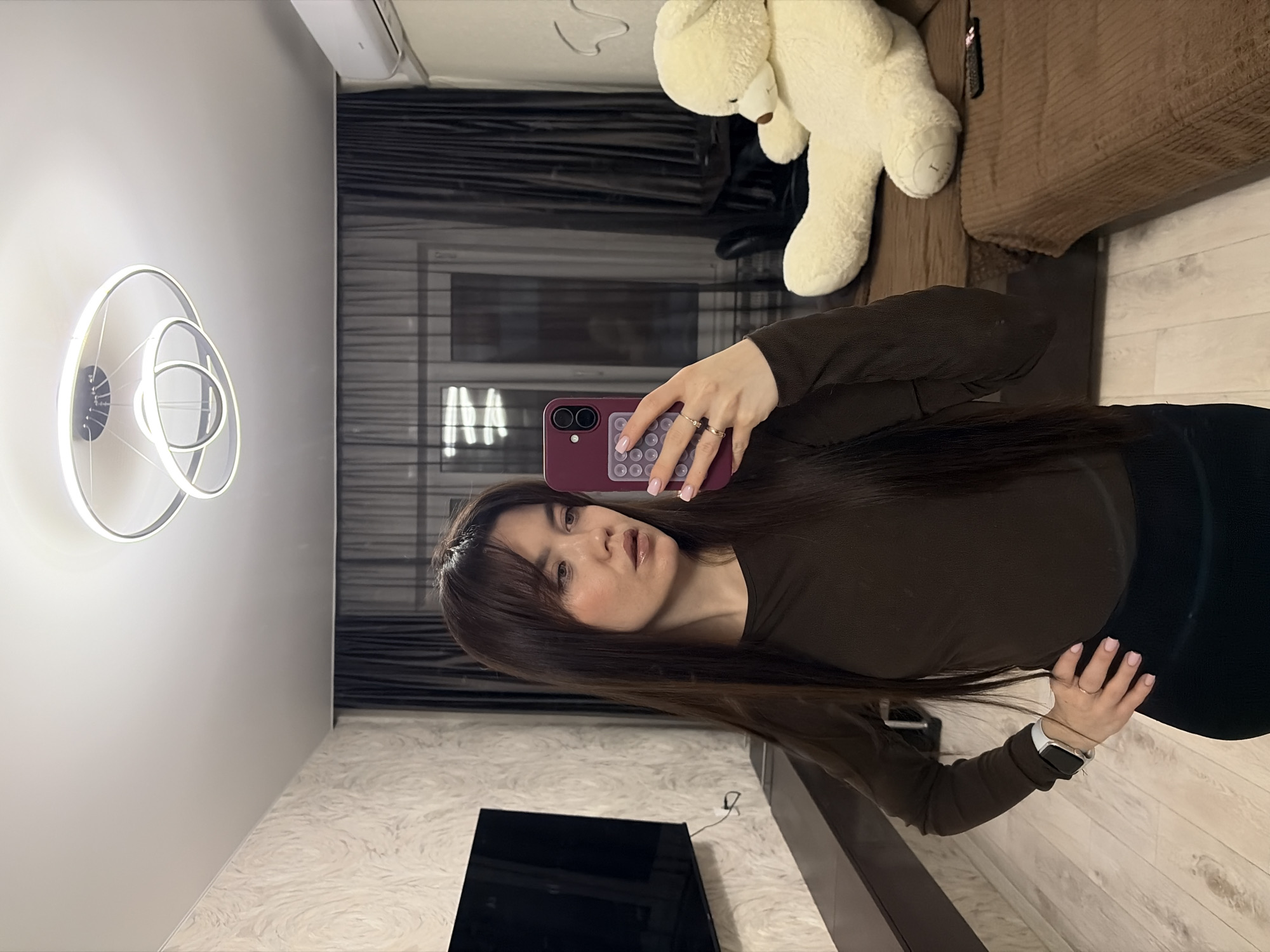 A brunette woman with bangs is standing indoors and holding a smartphone. The camera's flash is visible. She is wearing a brown long-sleeved shirt and black pants. She wears a smartwatch on her left wrist and a ring on her left hand. The background includes a wall-mounted decorative light fixture, a television, curtains, a teddy bear on a couch, and light-colored flooring. Based on the presence of common home furnishings and the fact that the photo appears to be a selfie taken by the woman, it is likely the photograph was taken in a residential setting, perhaps her home, in a city. The city is Votkinsk, Russia.