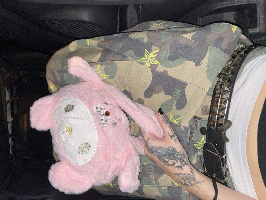 A person sits in a car with a pink stuffed rabbit on their lap, covered by camouflage-patterned clothing. The person's hand, adorned with tattoos, rests on the rabbit's ear. A studded belt is visible next to a white shirt. The scene is dimly lit.