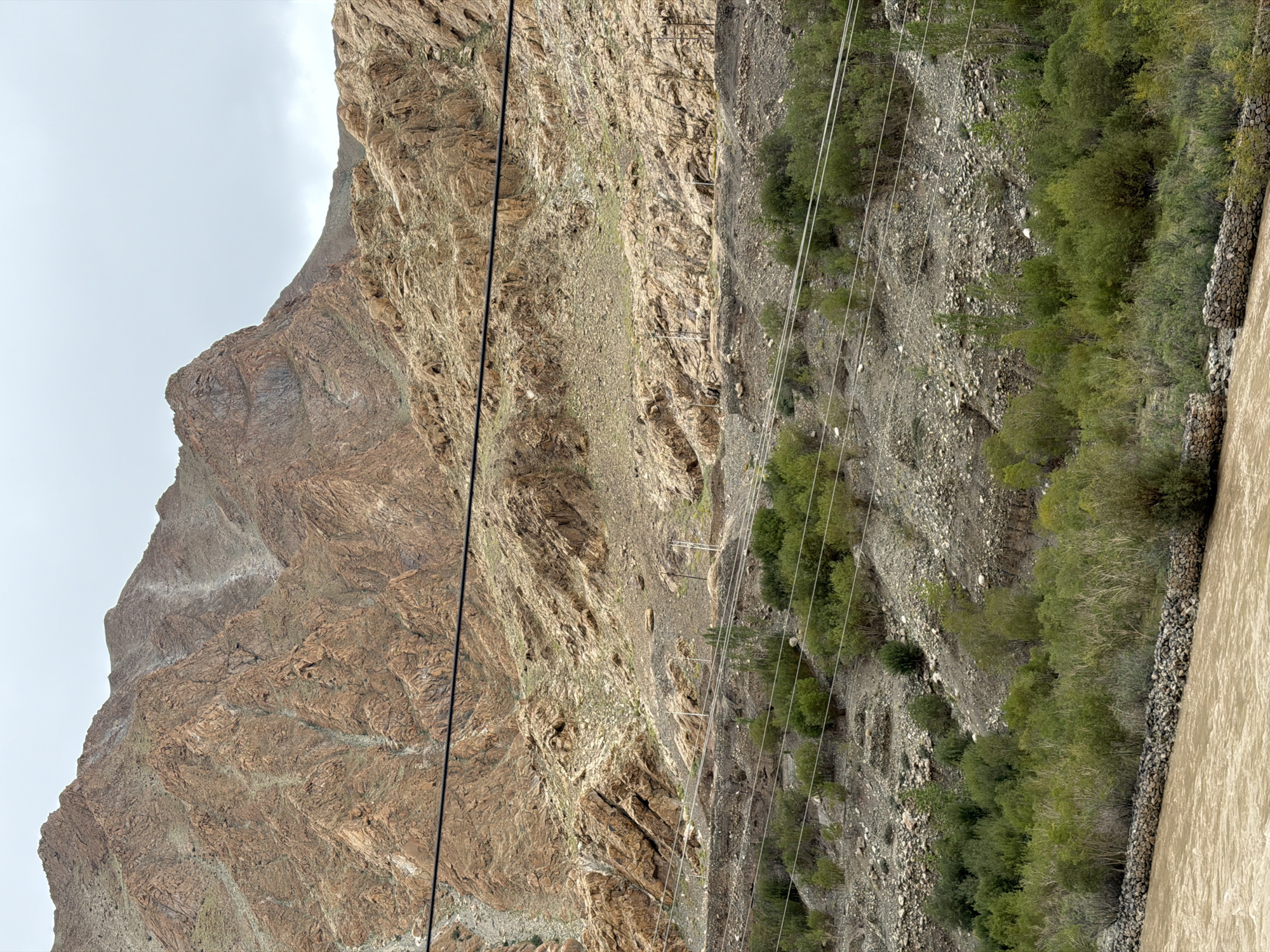 The image captures a wide shot of a mountainous landscape in Kungyamdo, India. The foreground features a wide, brown river, its banks reinforced with stone walls. Dense green vegetation, including trees and shrubs, lines the river. Further back, the terrain rises into rocky slopes dotted with smaller plants and debris. In the distance, massive, rugged mountains dominate the frame, their surfaces textured with rock formations and sparse vegetation. The sky is overcast with clouds. Power lines stretch across the middle ground, connecting to utility poles visible on the slopes. No people or animals are visible in the scene.