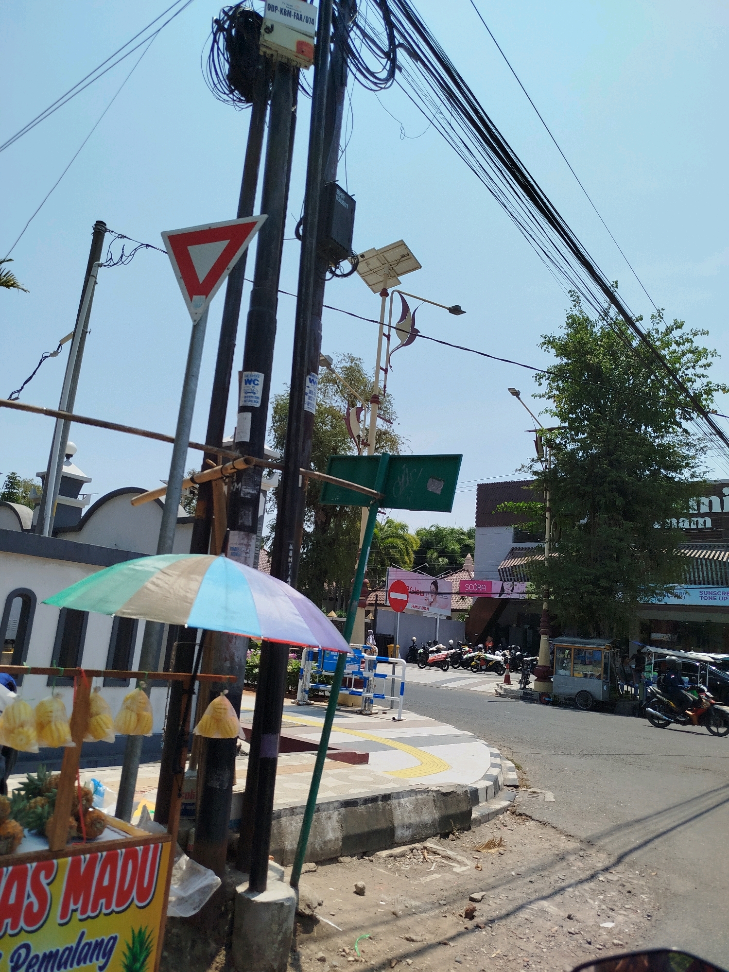 The image depicts a street scene in Kebumen, Indonesia, on a clear, sunny day. In the foreground, a small roadside stall displays pineapples and other produce under a colorful umbrella. The stall has signage reading "MAS MADU Pemalang". To the left, a white building with a domed roof and arched windows is visible, possibly a mosque.
The street is lined with utility poles adorned with numerous electrical wires, some appearing tangled. A yield sign and traffic signs are also present on the poles. Further down the street, a row of parked motorcycles and scooters suggests a commercial area. Several shops with awnings line the opposite side of the road, and a small food cart is visible. A person is riding a motorcycle on the road. The environment is urban, with pavement, a sidewalk, and some vegetation, including a large tree to the right. The overall impression is of a typical busy street in an Indonesian town.