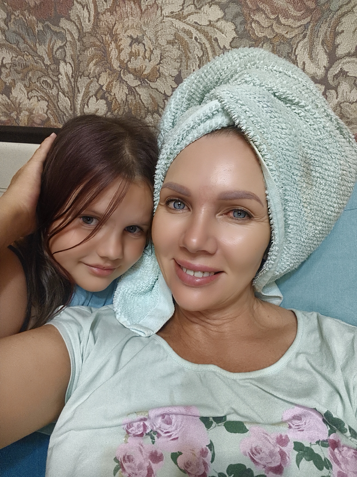 A medium shot shows two females, a child and an adult woman, posing for a selfie in an indoor setting. The woman, on the right, has blue eyes and is wearing a light blue towel wrapped around her head. She has a light skin tone and is smiling. The child, on the left, has brown hair and blue eyes and is looking towards the camera with a gentle smile. She is wearing a blue top, and her arm is resting on the woman's shoulder. The background features a patterned tapestry and a blue surface.