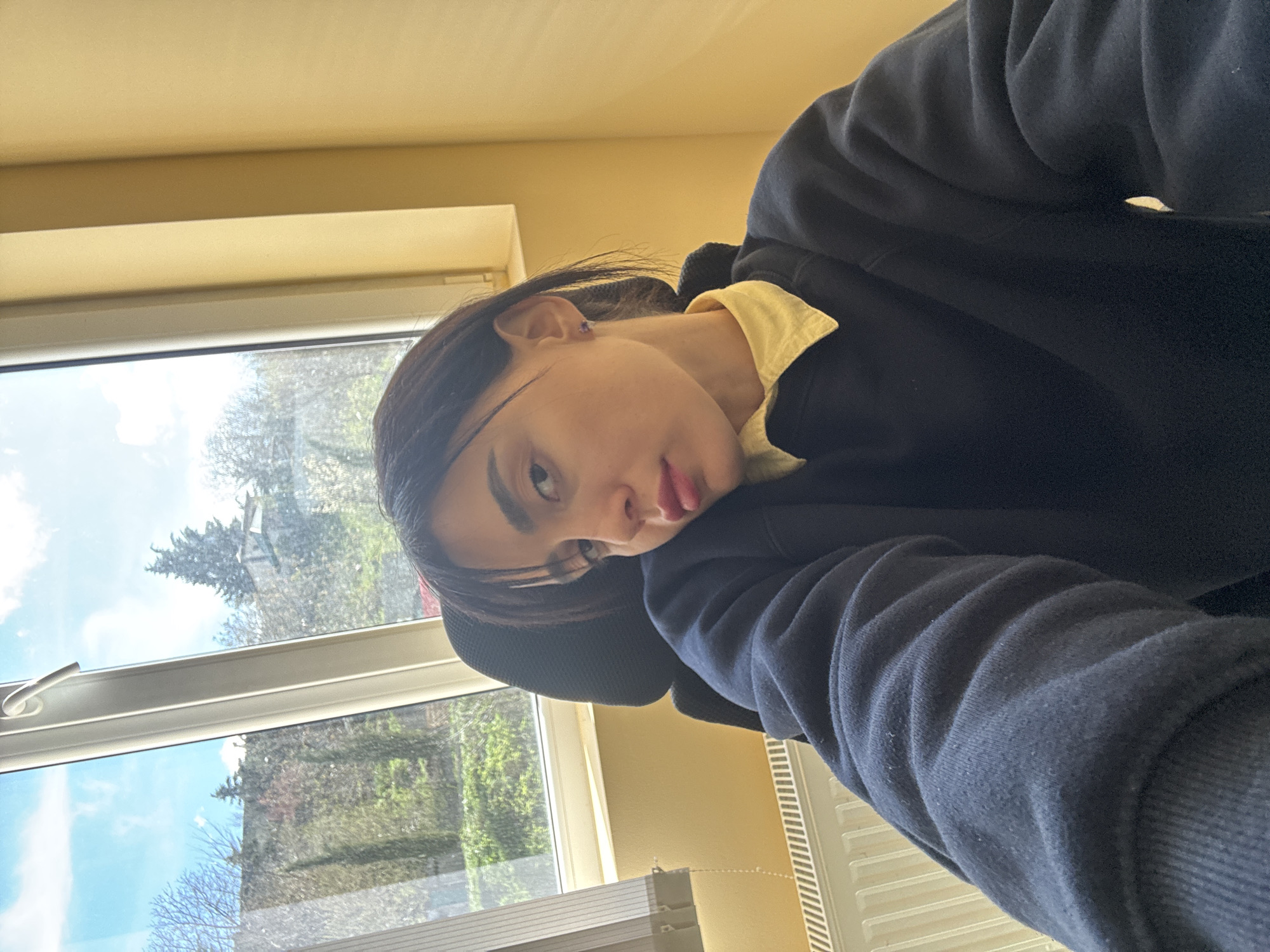 A young person is pictured in a selfie, positioned near a window. The subject has dark hair and is wearing a yellow shirt beneath a dark blue hooded sweatshirt. Outside the window, trees and buildings are visible, reflecting daylight. The setting appears to be indoors, with a radiator and office equipment partially visible. The location is likely Tbilisi, Georgia.