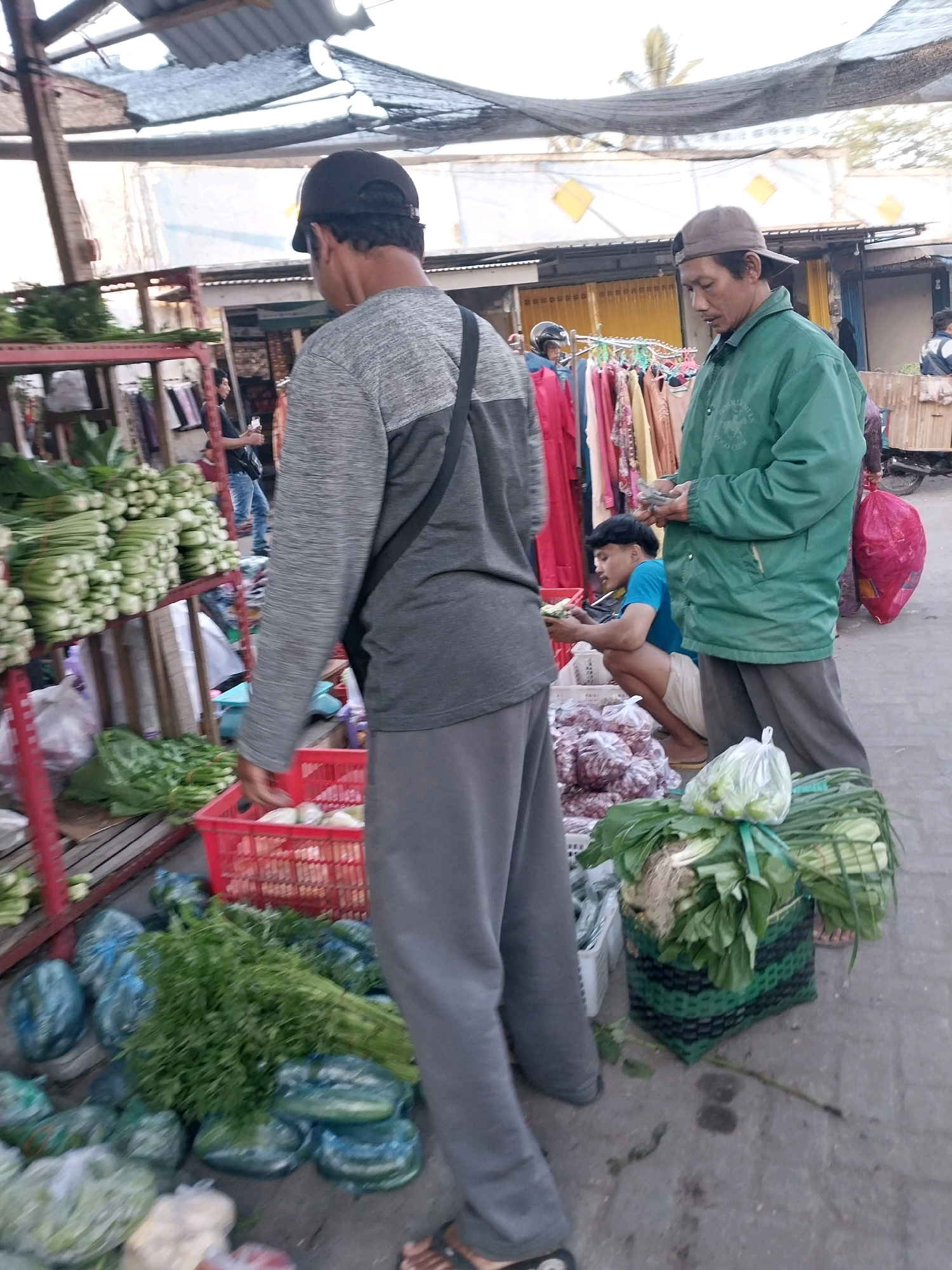 A man wearing a grey long-sleeved shirt and grey pants stands in the foreground of an outdoor market, facing away from the camera. He is holding a red basket containing produce. To his right, a man in a green jacket and grey pants stands facing left, holding some items in his hands. In the middle ground, a young man wearing a blue shirt and white shorts squats down, examining produce. Behind them, a rack of colorful clothing is visible. The environment is an outdoor market with stalls and various produce displayed. The overall scene depicts a typical market day.