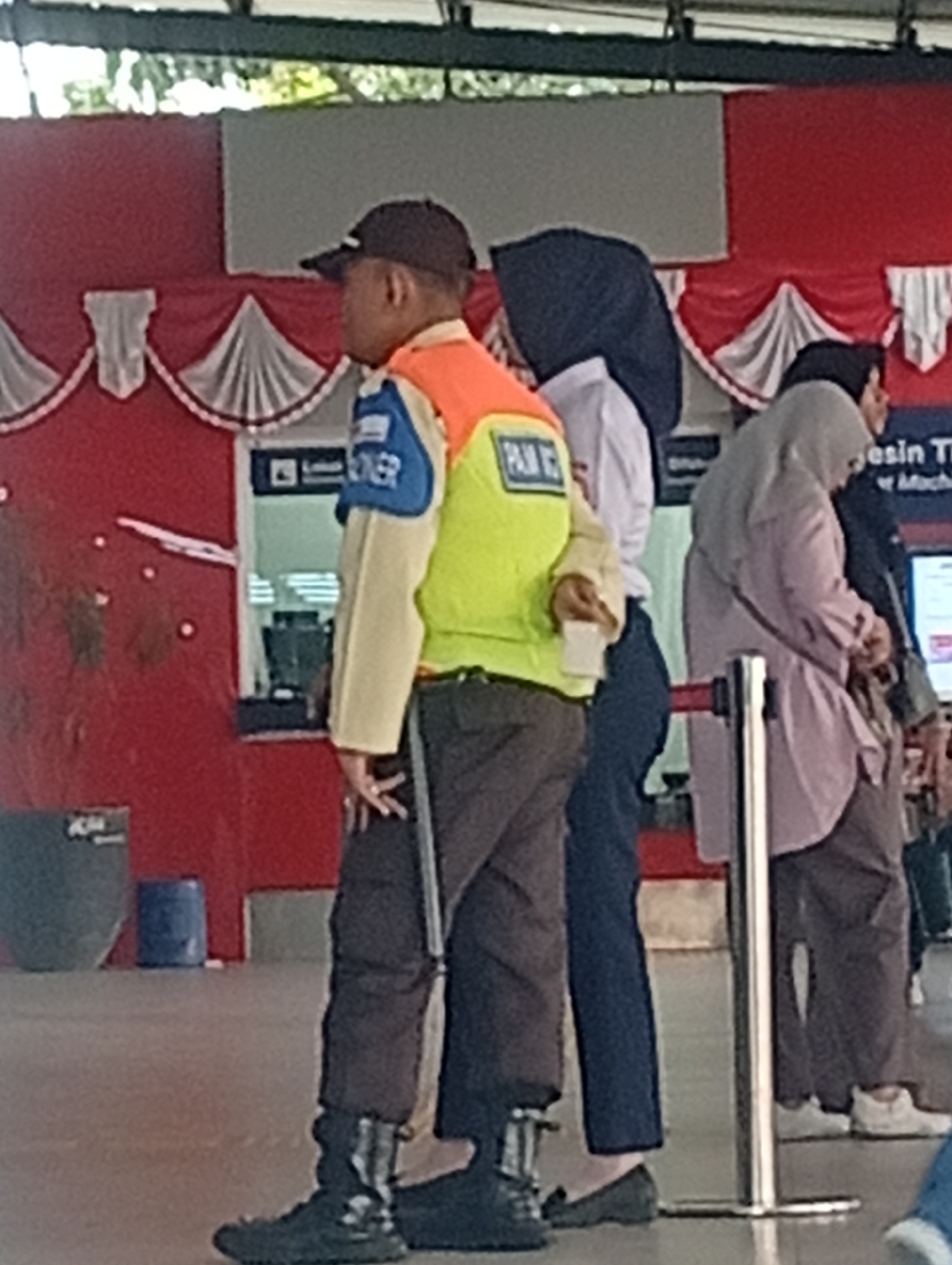 In Depok, Indonesia, a person wearing a black cap, an orange and green vest over a light-colored shirt, and dark pants stands holding a small white object in their right hand. This individual appears to be a security guard or staff member, indicated by the vest's markings which include the word "PAN". They are positioned in front of a red and white striped backdrop, possibly a decorative element at an event or facility. To their right, a person in a dark blue hijab and white shirt is partially visible, and further to the right, another person in a light grey hijab and a light purple jacket is also present. The environment suggests an indoor or semi-outdoor public space, with a metal stanchion in the foreground. In the background, there is a ticket booth or counter with a computer monitor visible inside.