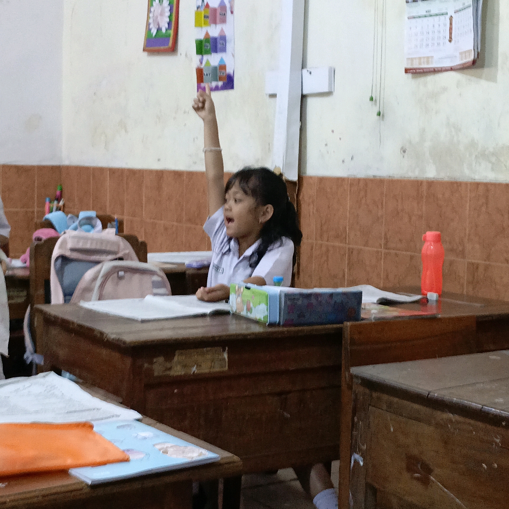 A young Indonesian girl in a white school uniform, with her hair pulled back in a ponytail, raises her right arm in a classroom setting in Salatiga, Indonesia. She is seated at a wooden desk, her mouth open as if speaking. On the desk, in front of her, are a book, a pencil case with cartoon characters, and a white eraser. To her right, on another desk, is a bright orange water bottle. In the background, the classroom walls are tiled with brown ceramic and painted white. Educational posters are visible on the walls, including a calendar. Backpacks and school supplies are stacked on chairs. The scene depicts a typical elementary school classroom environment.