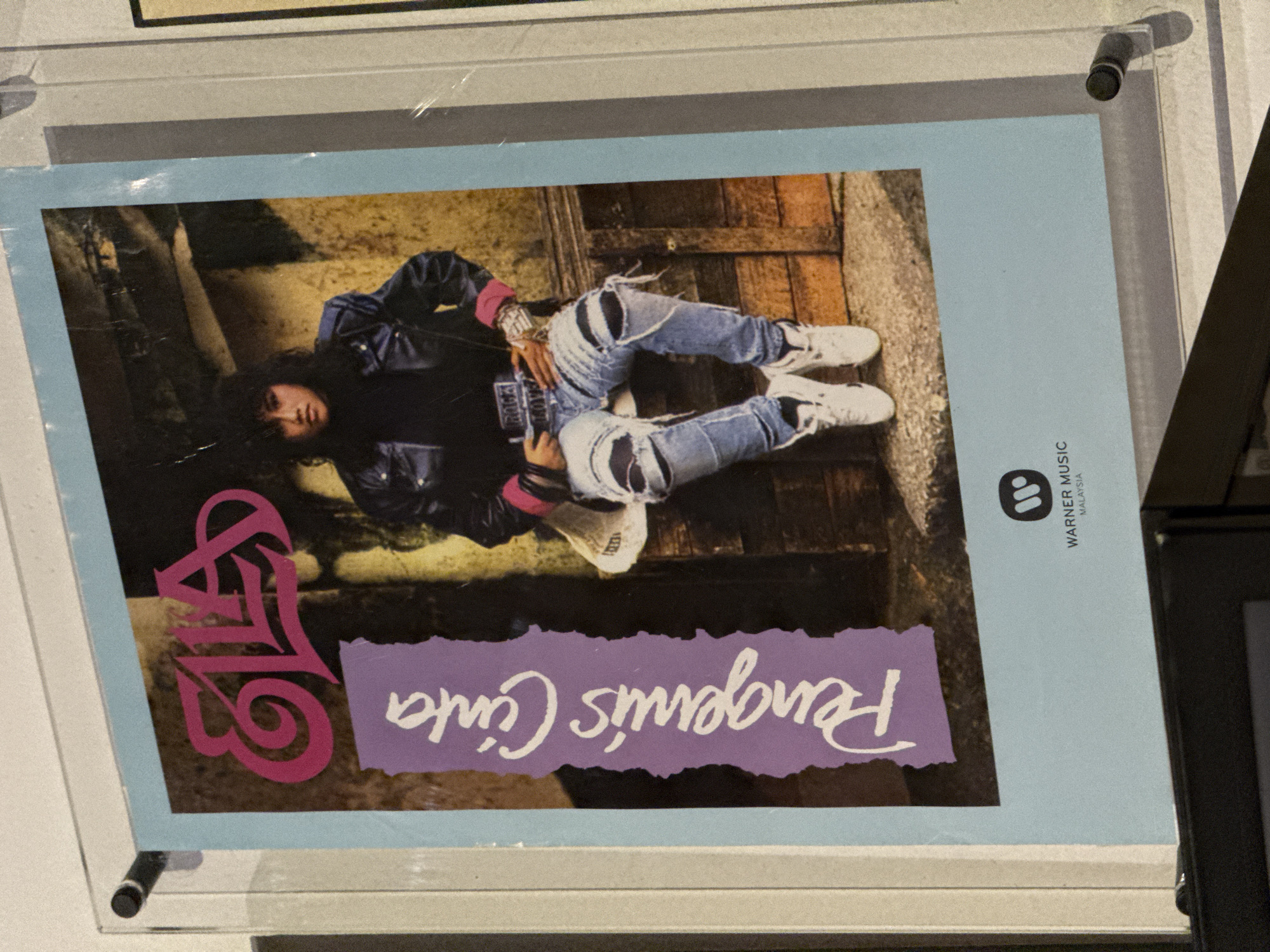 The image is a photograph of a framed album cover. The cover features a person lying down wearing a black jacket, pink shirt, distressed denim jeans and white sneakers. The person has dark hair. The album title, in script, appears to be “Pengemis Cinta”. A pink graphic design is located in the bottom left corner. The Warner Music Malaysia logo is in the upper right corner. The album cover is encased in a clear frame with standoffs. The context is an indoor environment. It is difficult to ascertain that the location is Kuala Lumpur, Malaysia without additional contextual clues.