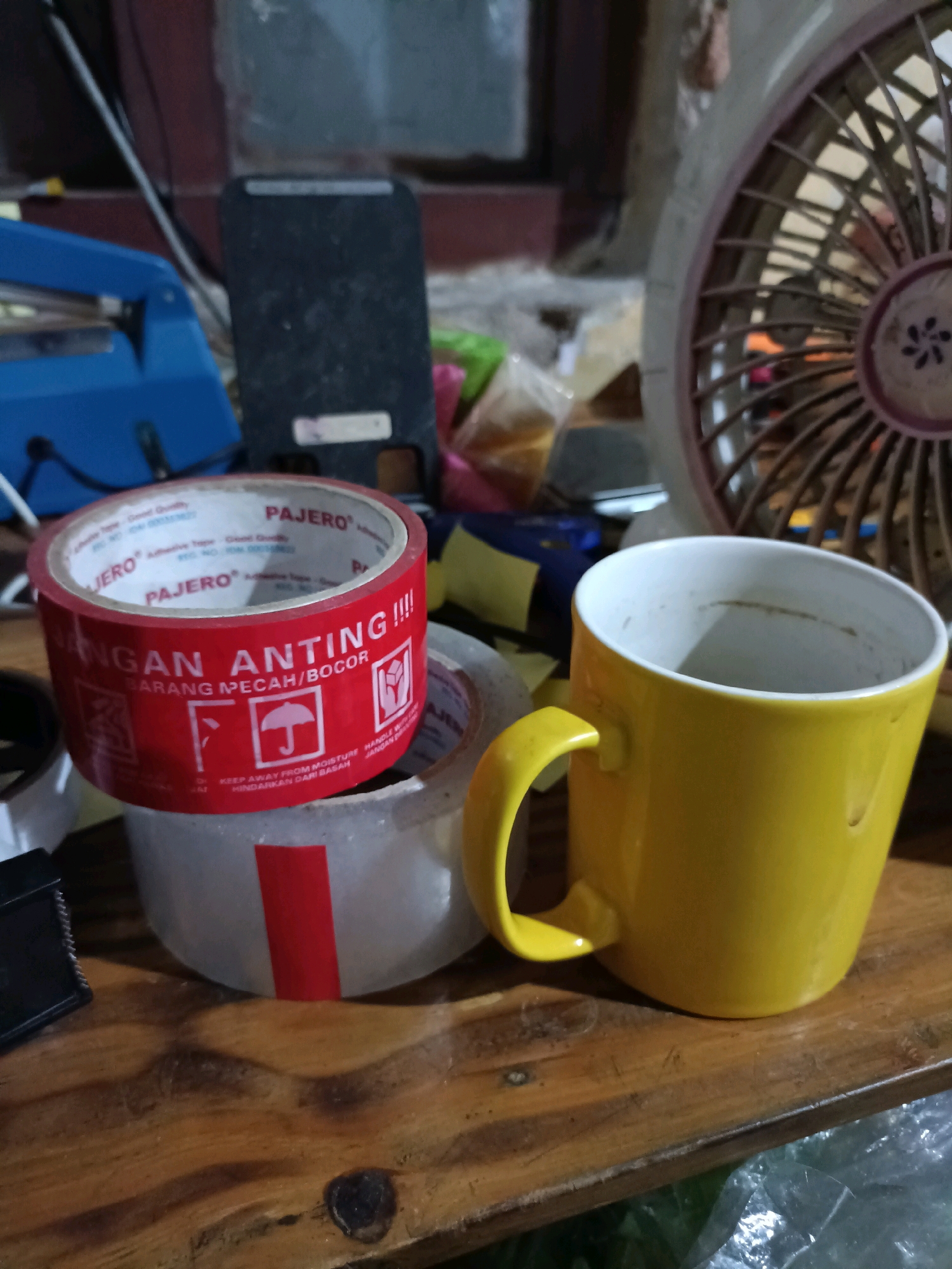 The image displays a close-up, eye-level, indoor shot of a cluttered wooden surface, possibly a table. In the foreground, a bright yellow mug with a white interior sits prominently on the right side. To its left, a roll of red packing tape with white lettering is stacked on top of another roll of clear tape that has a vertical red stripe. The red tape features warning symbols and text in Indonesian, including "JANGAN ANTING!!!! BARANG PECAH/BOCOR" (Do not shake!!!! Fragile/Leaking goods) and "KEEP AWAY FROM MOISTURE HINDARKAN DARI BASAH". Further to the left, out of focus, is a blue stapler-like object and a dark upright rectangular item. In the background, a portion of a brown and beige fan is visible on the right, and a blurry window or wall is present on the left. The lighting suggests a dimly lit indoor environment. The scene is devoid of any people or animals, and there are no discernible activities taking place. The overall impression is one of domestic clutter or a workspace. The location is identified as Ploso, Indonesia.