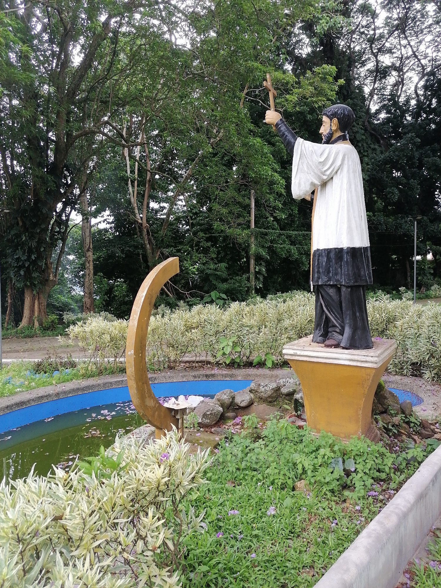 The image displays a statue of a bearded man, likely a priest or saint, standing in an outdoor garden or park setting in Davao City, Philippines. The statue is painted, depicting the figure in a white robe (surplice) over black clerical garments, with a wooden cross held aloft in his raised right hand. He faces away from the viewer, looking towards the right, embodying a solemn or blessing posture.
The statue rests on a yellowish, tapered pedestal positioned next to a small, circular pond. The pond has a blue-painted concrete rim, and its water appears green and murky, with scattered pink and purple flower petals floating on the surface. A distinctive golden-colored, crescent-shaped sculptural element rises from the water, possibly part of a fountain.
The environment is lush with vegetation, characteristic of a tropical climate. Tall, dense green trees form a canopy in the background and surrounding areas, their branches and foliage visible against the sky. The pond is bordered by light-colored, variegated bushes and various green ground cover plants, some with small purple flowers. The lighting suggests it is daytime, possibly under an overcast sky or during the softer light of morning or late afternoon, contributing to a peaceful atmosphere. There is no visible text in the image.