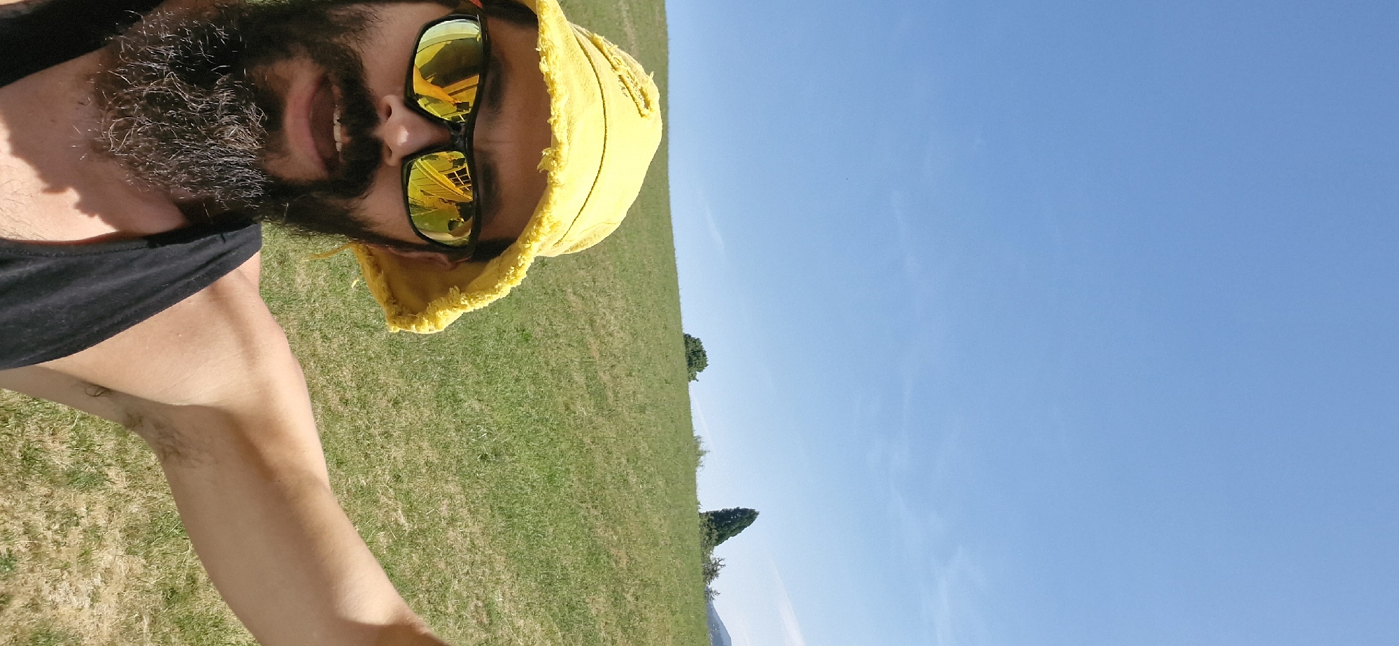 A man with a beard, wearing a yellow bucket hat and black tank top, is taking a selfie outdoors on a sunny day. He is wearing sunglasses that reflect a yellow and white structure, possibly a building or vehicle. The background consists of a grassy hillside under a clear blue sky. The location is identified as Wattenwil, Switzerland.