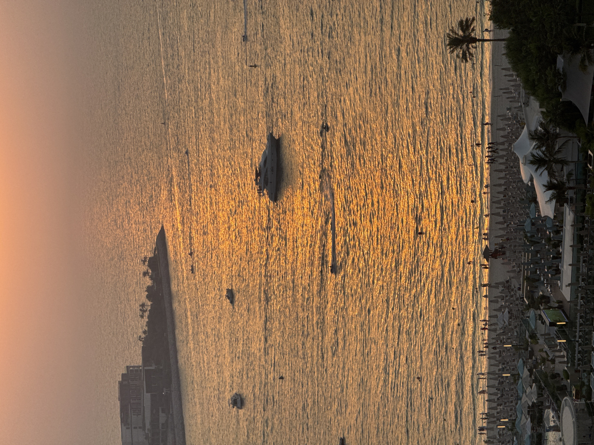 This image captures a sunset over the Arabian Gulf in Dubai, United Arab Emirates, likely from a high vantage point overlooking a beach resort. The water is a warm, golden hue, reflecting the setting sun which creates shimmering patterns on the surface.
In the distance, on the left side of the frame, is a modern hotel or resort complex situated on a peninsula. Several palm trees are visible along the coastline. The sea is dotted with various watercraft, including a large white yacht with people on its deck, several smaller boats, and what appear to be personal watercraft. Some individuals are also seen swimming or wading in the water.
Closer to the shore, a sandy beach is lined with rows of sun loungers and umbrellas, indicating a popular recreational area. Numerous people are strolling along the beach, and some are in the shallow water. The foreground reveals more of the resort's infrastructure, with more umbrellas, seating areas, and structures that suggest pools or open-air dining spaces. A lone palm tree stands prominently on the right side of the beach. The overall atmosphere is one of leisure and tranquility as the day draws to a close.