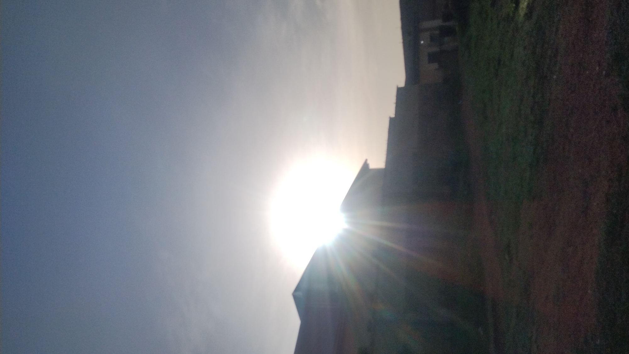 The image displays a dramatically backlit outdoor scene, likely captured during sunrise or sunset. The sun is positioned low in the sky, slightly to the right of the center, appearing as an intensely bright, overexposed sphere. Its powerful light causes significant lens flare, emanating outwards in distinct rays across the lower right quadrant of the sky. The sky transitions from a muted blue-grey in the upper left to a brilliant, almost white glow around the sun, indicating clear weather with some atmospheric haze.
Silhouetted against the bright sun on the right side of the frame are the dark, indistinct forms of multi-story buildings, their upper sections and rooflines sharply contrasted against the luminous background. Below these buildings, in the lower right, a dark, textured area suggests ground cover, possibly grass or earth, heavily shadowed and further obscured by the sun's glare. No people are visible, nor is any discernible text. The scene is characterized by its stark contrast between light and shadow, emphasizing the sun's intense brilliance over the obscured foreground elements in an unknown location.