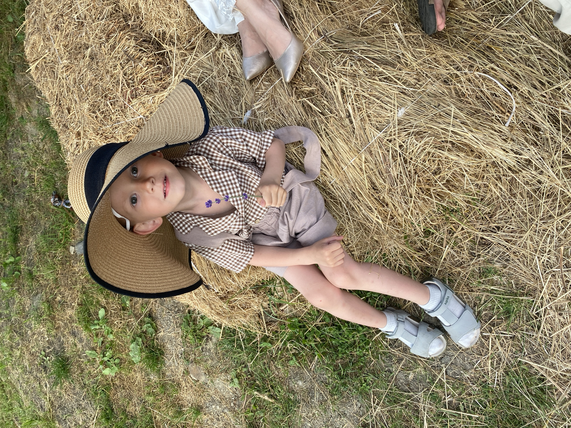 A young child lies on a haystack, wearing a large straw hat, a brown and white checkered shirt, and light brown shorts. The child has a few small purple flower decorations on their chest. They are wearing white socks and grey sandals. To the right of the child, two adults' legs are visible, one wearing silver heels and the other wearing dark shoes. The scene appears to be outdoors, likely in a rural setting, given the haystack and surrounding grass. The lighting suggests it could be daytime, possibly late afternoon. The child's expression is one of mild curiosity or surprise. The image is oriented vertically, with the child in the lower left quadrant and the adult legs in the upper right. The word "kapika" is visible on the child's sandals.