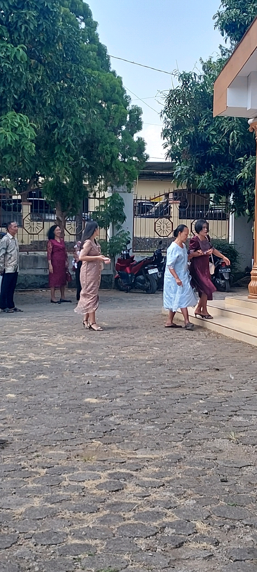 The image depicts a group of people outdoors, possibly at an event or gathering. The ground is paved with dark, irregularly shaped stones. In the background, there is a fence and vegetation, including trees with dense foliage. A red motorcycle is parked near the fence. Several people are visible, most of them women dressed in traditional or formal attire. Some are walking on the paved area, while others are ascending a set of stairs leading to a building with an orange-colored overhang. The lighting suggests it is daytime, likely with clear or partly cloudy skies. The scene appears to be set in Keling, Indonesia, indicated by the presence of traditional dress and architectural elements that might be characteristic of the region.