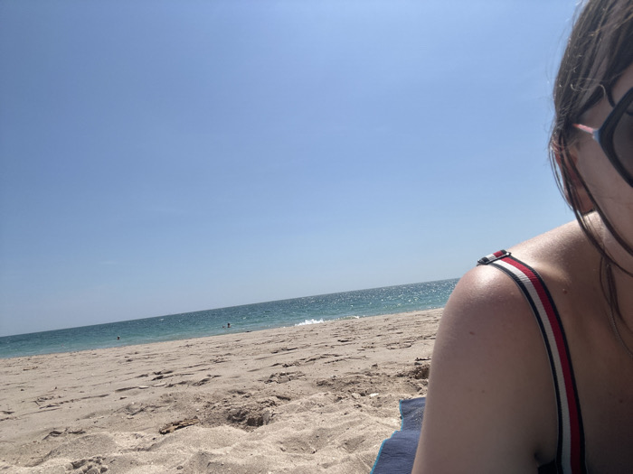 The image displays a beach scene in Phra Ae, Thailand, under a clear, bright blue sky. The foreground, positioned in the lower right, features the partial view of a person's left shoulder and the side of their head. The person wears dark-rimmed sunglasses and a swimsuit top with a red, white, and black striped strap over their shoulder. A corner of a dark blue towel or mat is visible beneath them on the sand. The midground consists of light brown sand, extending towards the water. In the background, the turquoise-blue ocean stretches horizontally, with small waves visible at the shore. Several indistinct figures, likely people, are visible in the water. The upper half of the image is dominated by a cloudless, light blue sky.