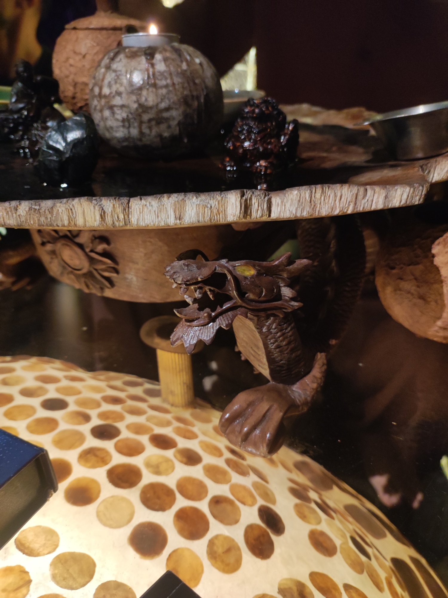 A wooden sculpture of a dragon with green eyes is positioned on a wooden table under an ornate wooden platform. The platform holds several decorative items including a light-colored, round candle holder with a lit candle, a dark round object, and an urn-like item. The dragon sculpture rests on a small, cylindrical wooden stand. Underneath the dragon is a lamp with a circular pattern of brown spots on a light background. The location in the city of Lokaserena, Indonesia cannot be determined from the image.