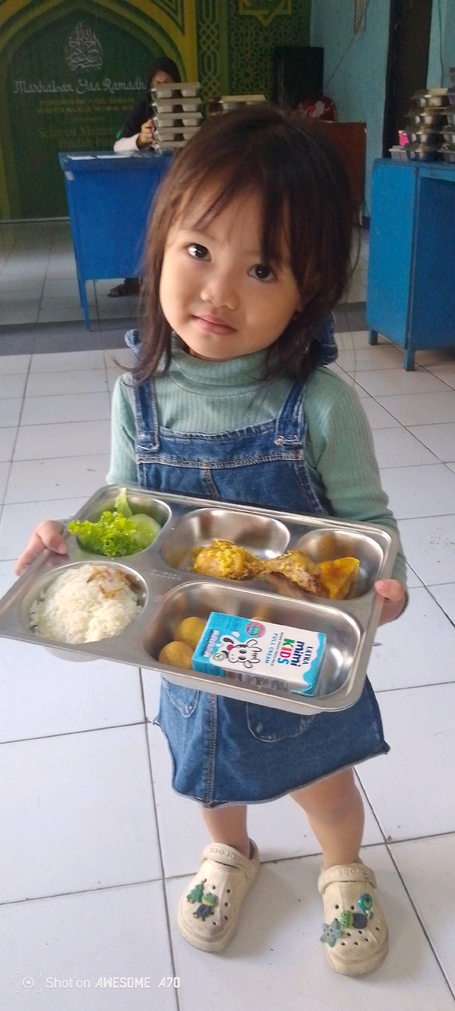 A young Indonesian girl, likely between 2-4 years old, stands in an indoor setting and holds a metal tray of food. She has dark hair and is wearing a light blue long-sleeved shirt and a denim overall dress. The tray contains rice, a salad of lettuce and cucumber, two pieces of fried chicken, small meatballs, and a small carton of "Ultra Mini Kids" full cream milk. The girl is wearing beige Crocs-style shoes adorned with charms. The background suggests a school or community center, possibly a cafeteria, with blue furniture and a green wall decoration with Islamic calligraphy and patterns. A stack of trays is visible on a counter in the background. The floor is tiled. The image appears to be taken in Bandung City, Indonesia, based on the visible timestamp "Shot on AWESOME A70" which is often associated with Samsung phones popular in the region.