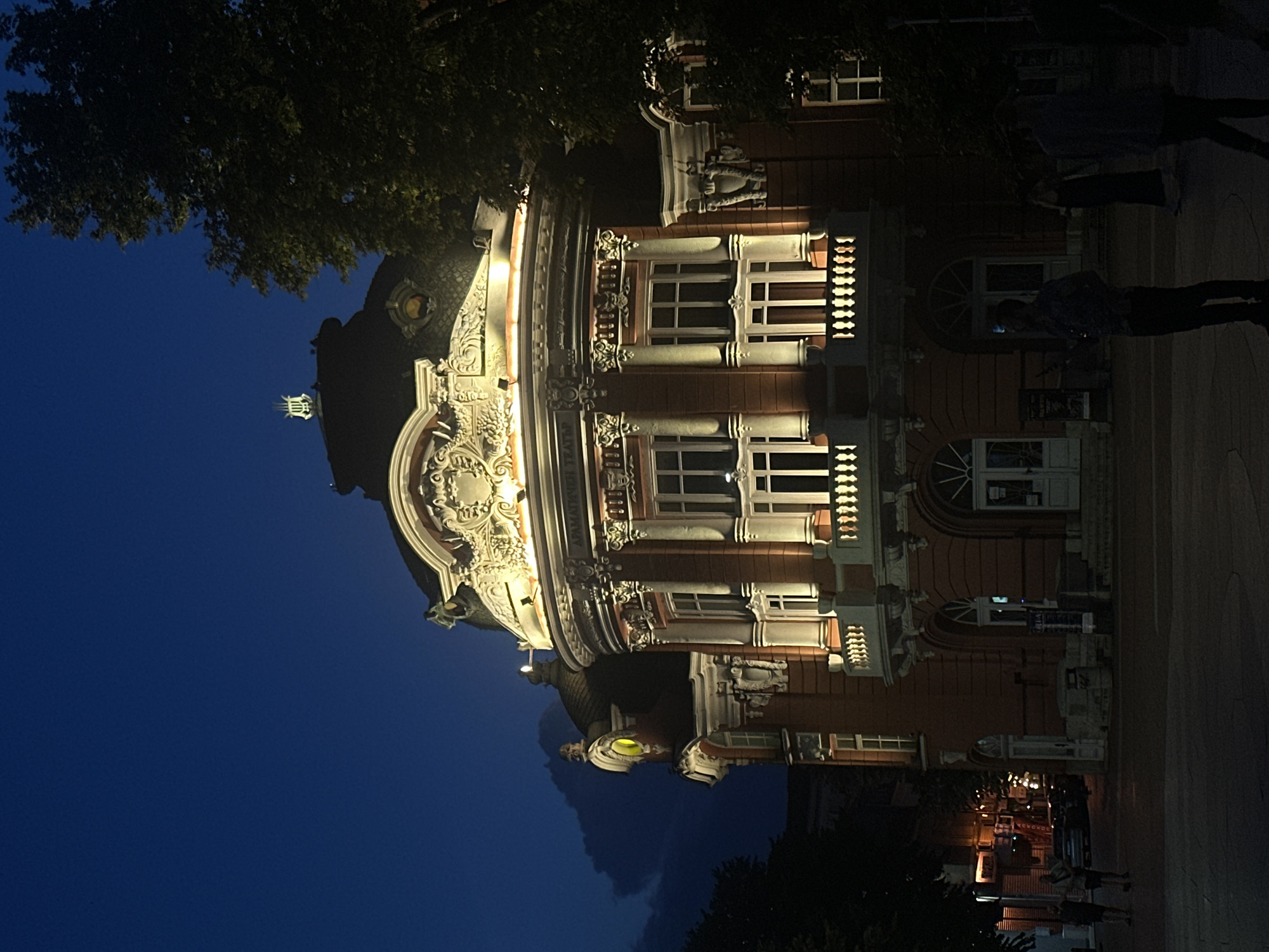 The image shows a grand, ornate building lit up at night, likely a theater or opera house, with a dark blue sky above. The building is made of red brick and features intricate white architectural details, including columns, balconies, and decorative carvings. Lights highlight the upper floors and roofline. A large tree with dark foliage partially obscures the right side of the building. In the foreground, a few silhouetted figures can be seen standing on a paved plaza, some looking towards the building. The overall atmosphere is one of evening twilight in a city setting. The building has a prominent lyre-shaped finial on its roof.