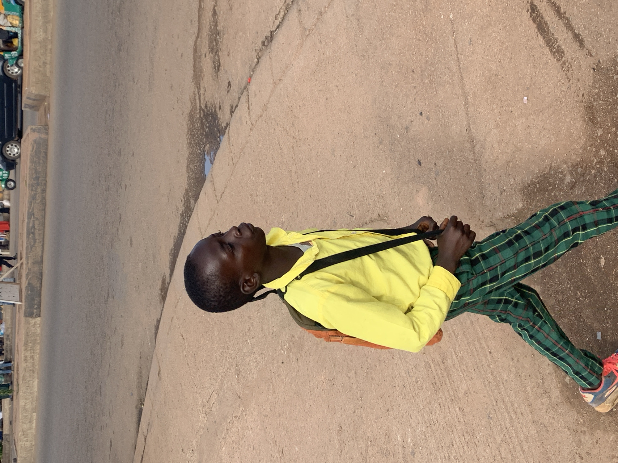 A young boy wearing a bright yellow long-sleeved shirt and green plaid pants walks along a paved sidewalk in Gura Top, Nigeria. He has a small orange backpack slung over his shoulders. The sidewalk is cracked and shows signs of wear, with some water puddles on the adjacent asphalt road. In the background, several vehicles, including a black car and a three-wheeled auto-rickshaw, are visible on the road. Other pedestrians can be seen in the distance, some standing and others walking. The scene appears to be set during daylight hours.