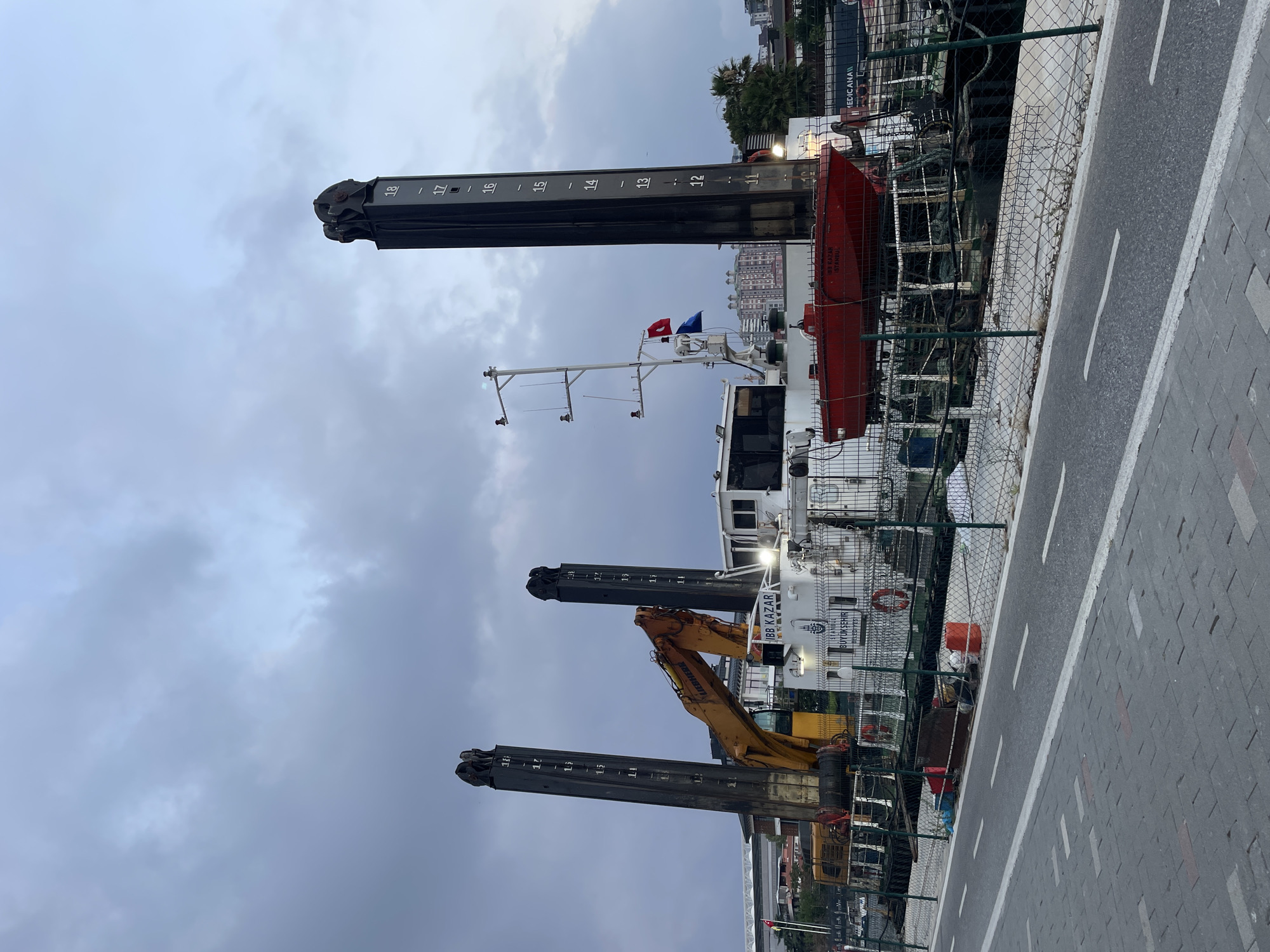 The image depicts a large, industrial barge docked at a harbor, likely in Kadıköy, Turkey. The barge is equipped with two prominent, tall, black vertical structures, possibly for lifting or guiding heavy objects. These structures have numerical markings indicating depth or height. An orange excavator, manufactured by Liebherr, is positioned on the deck of the barge. The barge itself is white with the inscription "IBB KAZAR ISTANBUL" and the Istanbul Metropolitan Municipality logo, suggesting it is owned or operated by the municipality. A bright red lifeboat is mounted on the deck. The surrounding environment includes a chain-link fence, a paved walkway with white lane markings, and a cloudy sky. In the background, buildings and possibly a stadium or large structure are visible, consistent with an urban waterfront setting. No people are visible in the image. The scene appears to be a static view of port infrastructure, possibly used for construction or maintenance work.