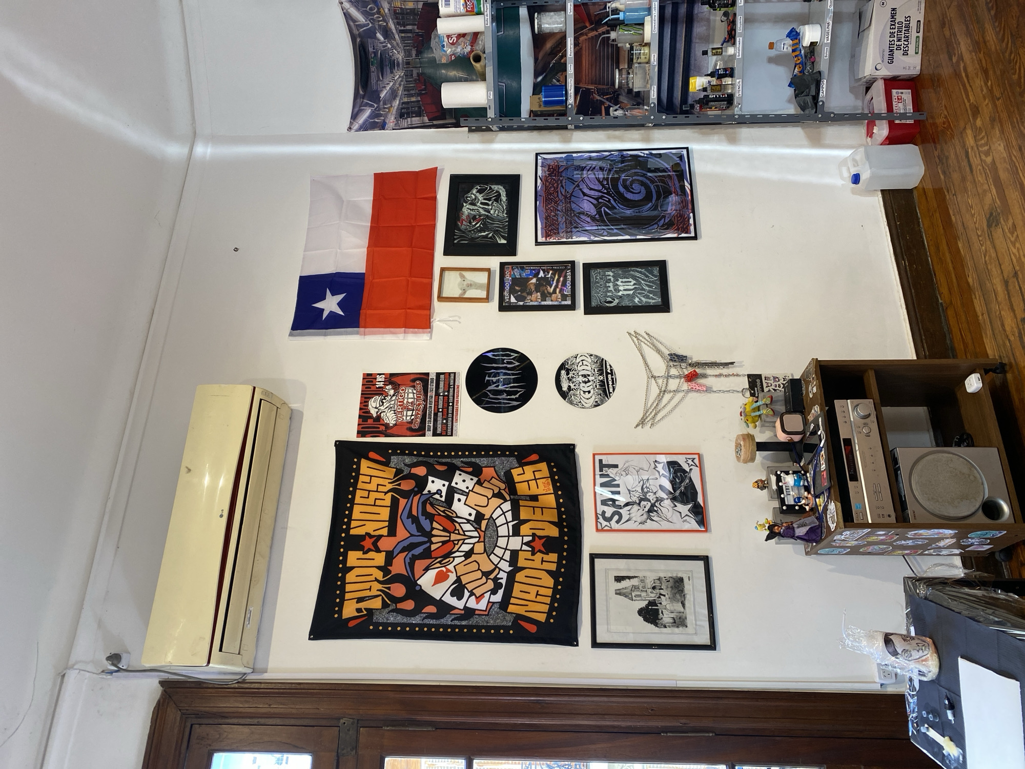 The image displays an interior wall in what appears to be a creative workspace or studio, likely in Buenos Aires, Argentina, given the context. The wall is densely decorated with various items, from practical supplies to personal art and memorabilia.
At the top, a metal shelving unit spans the wall, filled with numerous bottles, rolls of paper, and containers. Labels visible on these items include "Diluyente" (thinner), "STENCIL," "Bolsas" (bags), "VASELINA" (vaseline), "CAMPOS," and "alcohol," strongly suggesting tattoo or art supplies. On the wooden floor below, a box of "GUANTES DE EXAMEN DE NITRILO DESCARTABLES" (disposable nitrile examination gloves) in size "M" is visible, alongside a white plastic jug.
The main wall area is adorned with a variety of framed artworks and decorations. A Texas flag hangs prominently on the left. Nearby, framed pictures include a dark, abstract piece with purple swirls, another with a skull motif, and a black and white drawing of a house. A large black fabric banner featuring stylized hands, dice, and playing card suits with text "TUOC NOSSO" and "NADE DELES" is a central decoration. Smaller circular black decorative items are also displayed.
A poster or flyer on the wall details an event, with text such as "VERTIGO," "14 HS," "2357STUDIO," "DROP COLABORACIÓN VERTIGO 2037 STUDIO," "DI SET FLASH TATTOO," "SERIGRAFIA EN VIVO CLOTHING & VINTAGE," and "ENTRADAS POR PASSLINE." Other smaller framed pieces also show "VERTIGO" and "VERTIGO GO."
To the left, an LG air conditioner unit is mounted near the ceiling. On the right, a wooden shelf holds electronic equipment, specifically a silver stereo receiver and a speaker. This shelf is also decorated with small figurines, including one wearing a witch's hat, and a smartwatch. The room features a wooden plank floor and warm indoor lighting.
