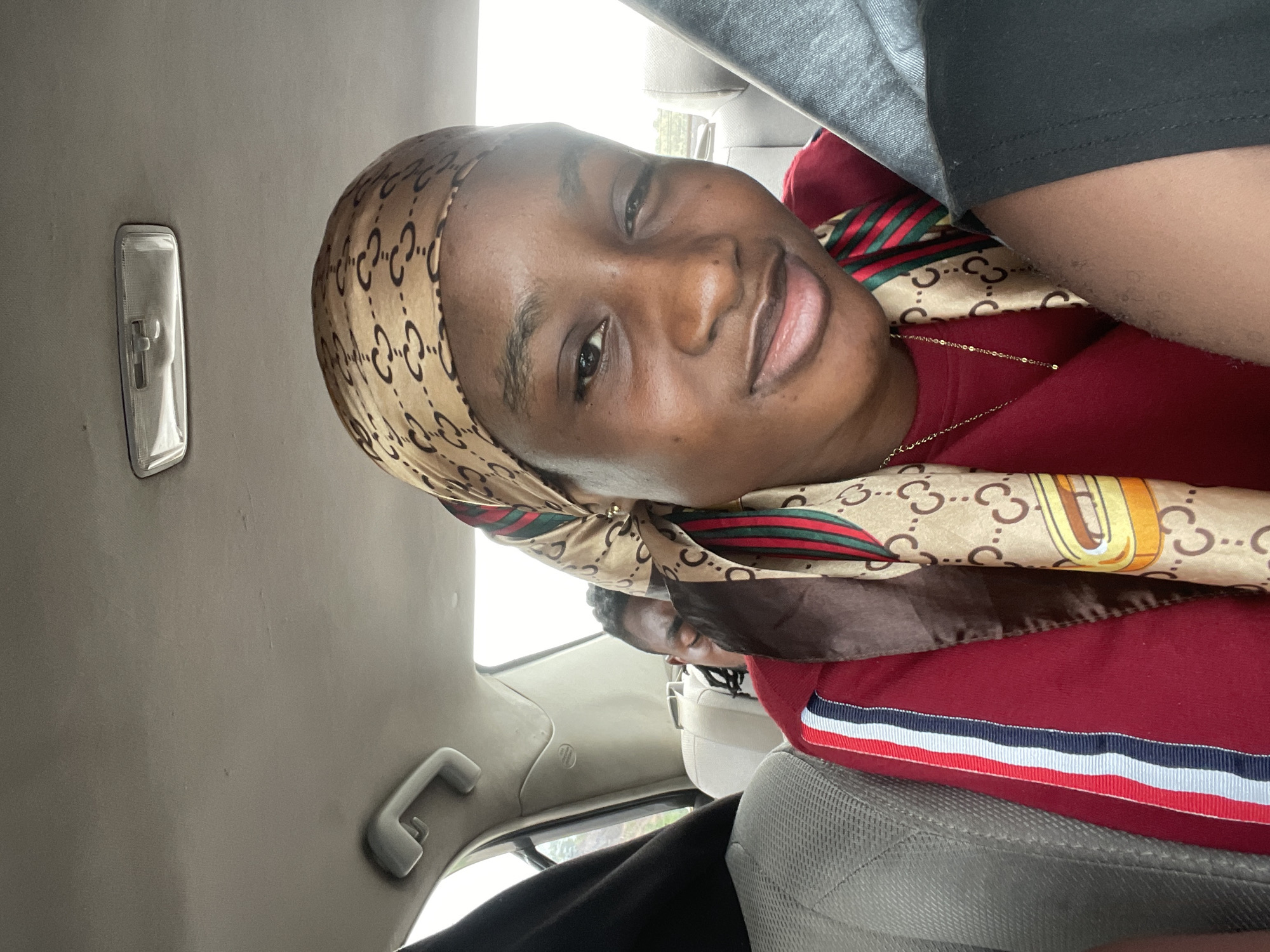 The media file shows a close-up of a young Black woman inside a vehicle. She is wearing a headscarf with a repeating logo pattern and a red long-sleeved shirt with striped trim. The woman is smiling and looking towards the camera. The interior of the vehicle is visible, including a gray ceiling with a light fixture and a grab handle. Another person is partially visible in the background, also inside the vehicle. The lighting suggests it is daytime, possibly overcast. The overall scene appears to be a casual moment within a car, likely in Kwali, Nigeria, as indicated by the provided location context. There is no visible text in the image.