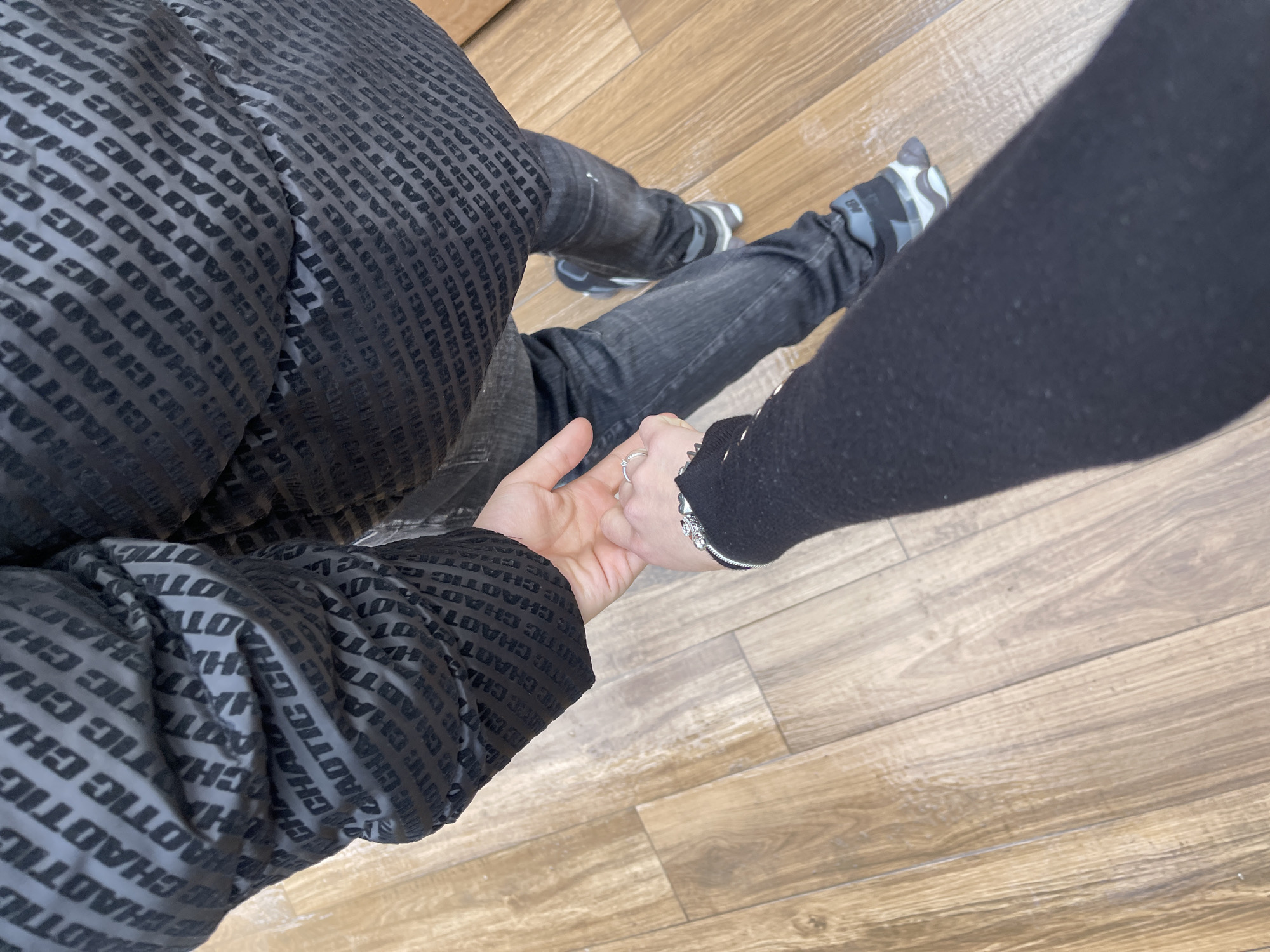 A high-angle shot shows two individuals holding hands against a backdrop of light-brown wood flooring. A person wearing black jeans, black shoes with gray and white trim, and socks with a gray and black pattern is visible. They are also wearing a black puffy jacket with "CHAOTIC" printed repeatedly on it. Intertwined with their hand is another person, who is dressed in a long-sleeved black shirt. A silver ring adorns one of the fingers, and a silver charm bracelet is visible on the wrist. No information is available to determine the location in Veliko Tarnovo, Bulgaria.