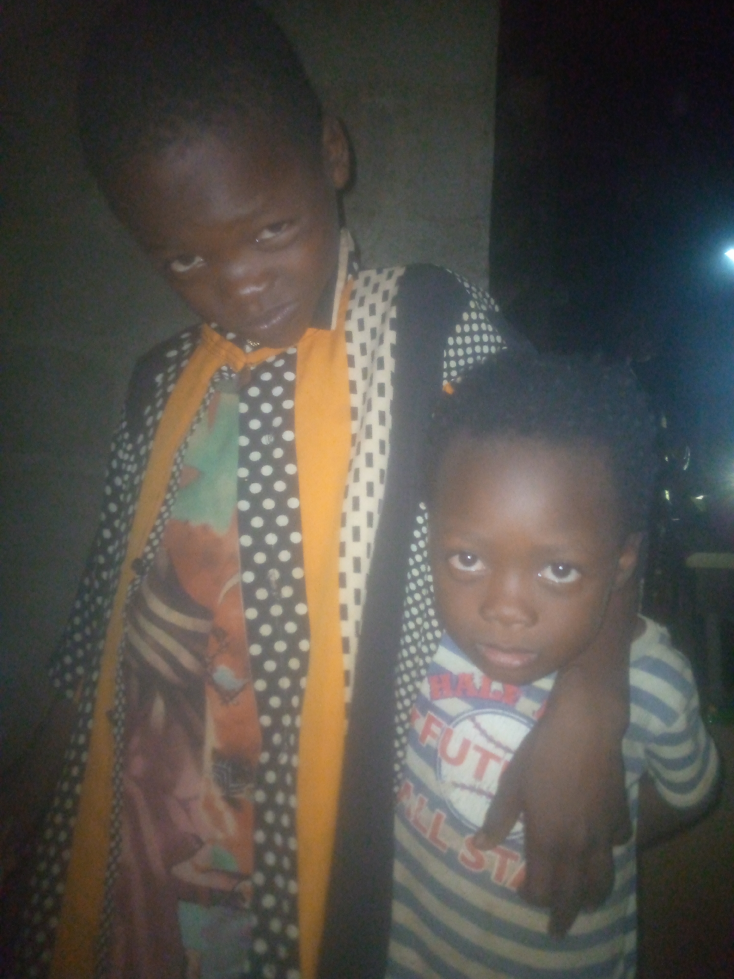 Two children stand close together, one slightly behind the other. The child in the front wears a striped t-shirt with the words "FUTURE ALL STAR" printed on it. The child behind has an arm around the shoulder of the child in front. The child behind wears a patterned shirt with multiple designs including polka dots, stripes, and blocks of color. The location appears to be indoors, with a wall or curtain visible in the background. The lighting in the scene is dim, making it difficult to discern finer details.