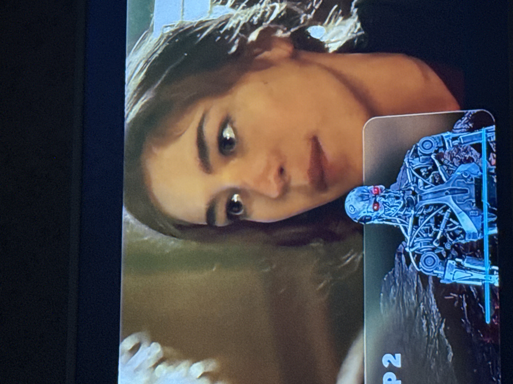 The image shows a television screen displaying a scene with a young woman and an overlay. The woman is the primary subject, with her face visible and looking towards the viewer. An overlay featuring a metallic robotic figure, possibly a Terminator, is present. The overlay also includes the text "P2." The scene appears to be a paused or still image from a video. The image does not contain location information about the city of Mytishchi, Russia.