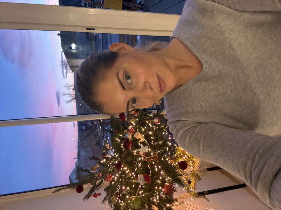 A selfie captures a woman indoors, positioned slightly to the right of the frame, with a decorated Christmas tree to her left and a large window in the background. The woman has her hair pulled back and wears a light grey V-neck top. She looks directly at the camera with a neutral to slight smile.
The Christmas tree is brightly lit with numerous warm white lights and adorned with a variety of ornaments, including red baubles, white and gold decorations, a small Santa figure, and star-shaped ornaments. Through the window, the sky displays soft hues of blue, purple, and pink, indicating either dawn or dusk. Distant outdoor elements, including what appear to be structures and bare trees, are visible against the colorful sky. The overall scene suggests a festive, cozy atmosphere, likely during the evening due to the illuminated tree.
The image is rotated 90 degrees clockwise, with the woman's head tilted to her left. This scene is set in Rousset, France. There is no visible text in the image.