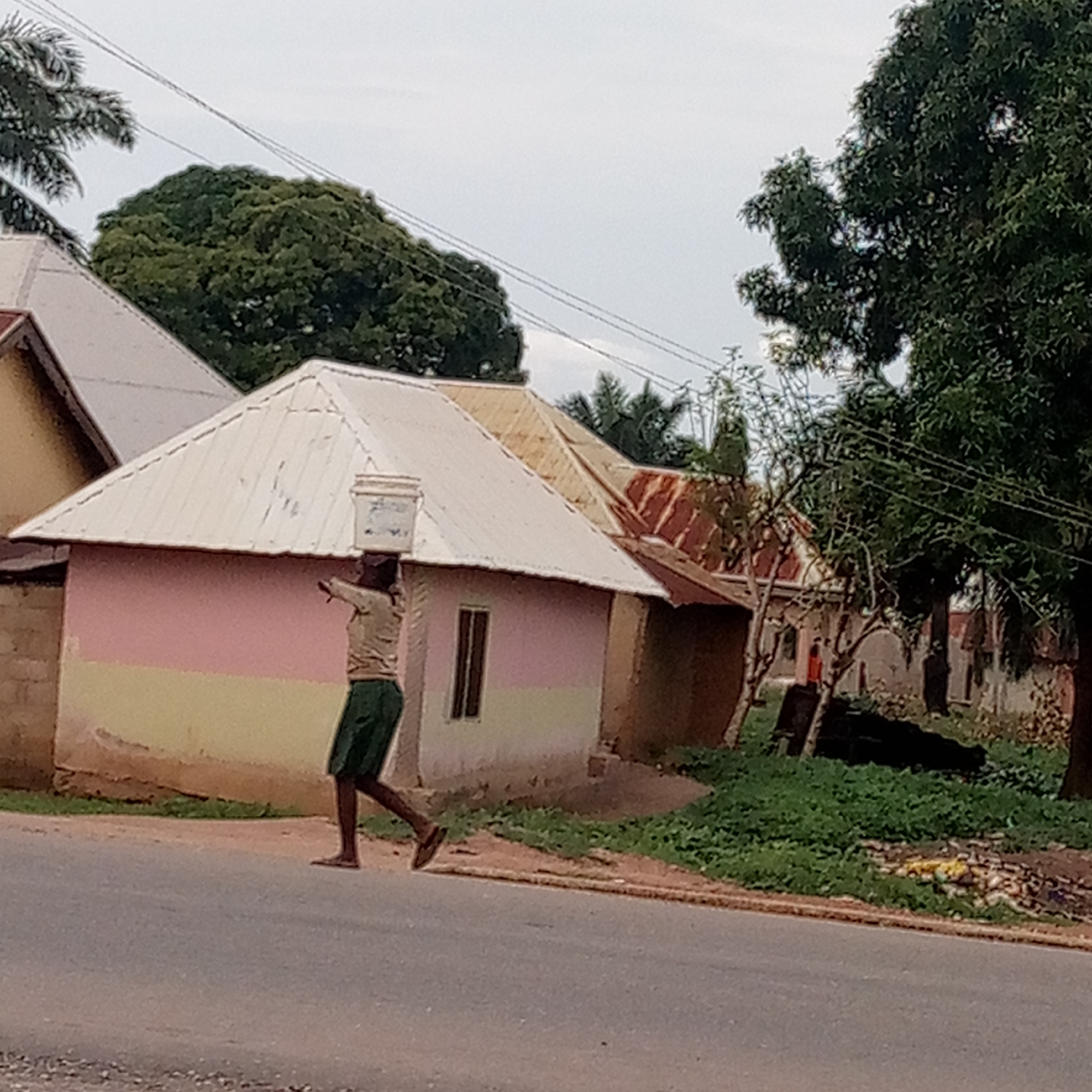 A street scene in Mararaba, Nigeria depicts a man walking along a paved road, carrying a white bucket on his head. He wears a tan shirt and green shorts. Behind him, a building is painted in shades of pink and yellow, with a corrugated metal roof. Nearby, other buildings with deteriorating exteriors and rust-colored roofing are visible. Trees and sparse vegetation are adjacent to the road. Overhead, power lines stretch across the sky, which is overcast. Litter is visible on the side of the road.